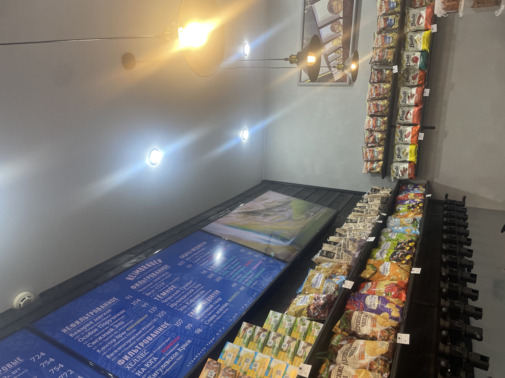 The image shows the interior of a store, likely a craft beer or beverage shop, with shelves stocked with various products and digital menu boards. One menu board displays text in Cyrillic, listing items like "НЕФИЛЬТРОВАННОЕ," "БАВАРИЯ МЯГКОЕ," and various types of beer and cider, along with prices. Another menu board features an image of a landscape. Below the menu boards, shelves are filled with packaged snacks, including bags of chips and other food items, also with Cyrillic labeling. The store has a modern industrial aesthetic with exposed lighting fixtures and dark shelving units. The presence of Cyrillic text and the likely product selection suggests the location is in a Russian-speaking country. There are no people visible in the image. The overall environment is well-lit, with both overhead lighting and task lighting from the menu boards. The store appears to be a retail establishment selling drinks and snacks.