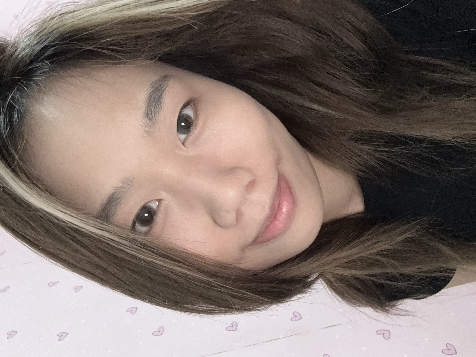 The image shows a close-up selfie of a woman with brown hair and blonde highlights. She is lying on a pink surface with a pattern of white dots and light purple hearts. The woman has fair skin, brown eyes, and pink lips. She appears to be wearing a black top. The background is out of focus, providing no clues to the city or the district.