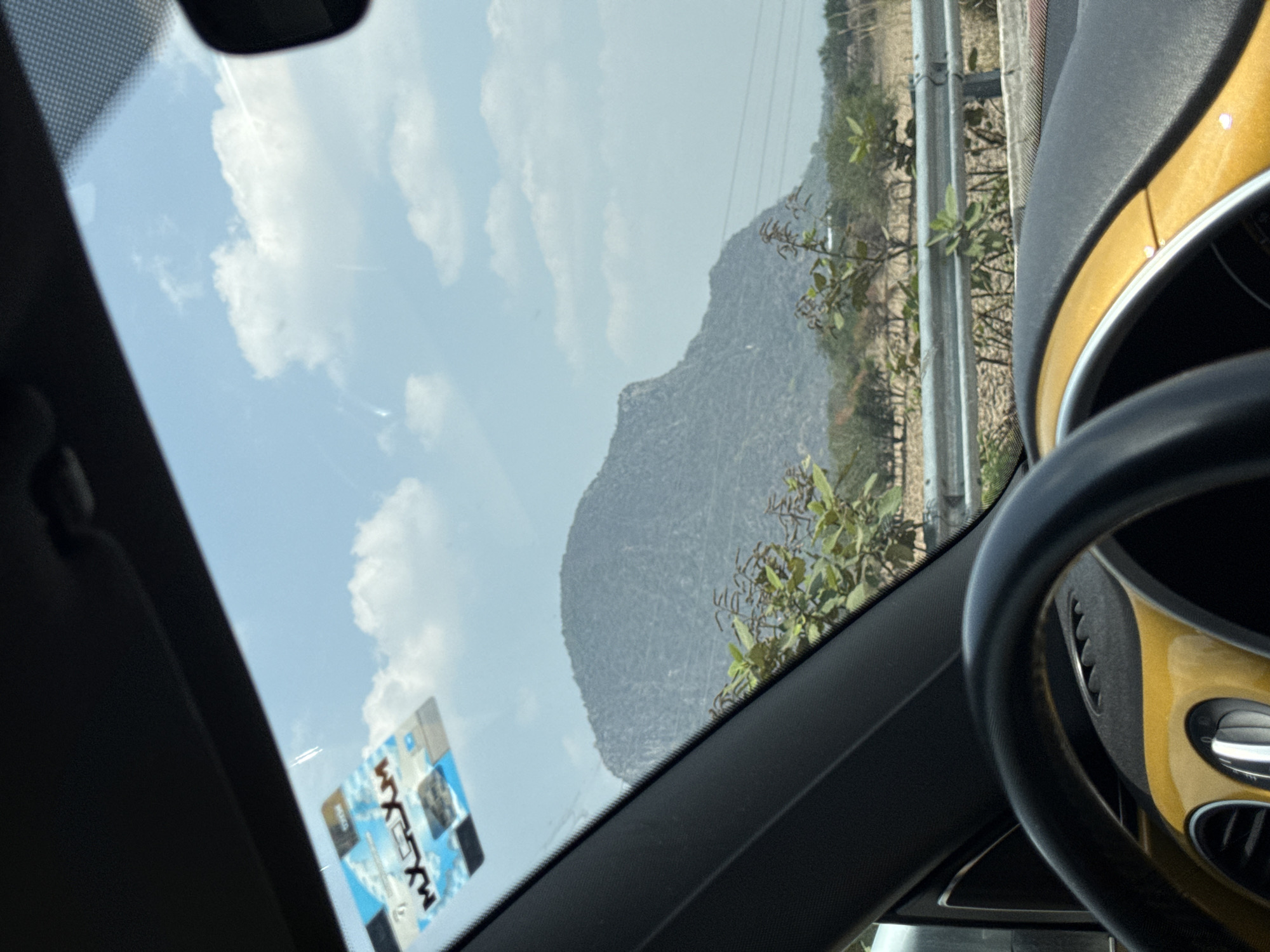 The image, taken from inside a car, shows a view through the windshield. In the background is a mountain, possibly viewed from the car travelling along a road, indicated by the guardrail visible. The sky is partly cloudy. In the foreground, the car's dashboard and steering wheel are visible. A permit sticker is on the windshield, with "MX CDMX" visible. The image suggests a location near a mountain in a rural or semi-rural setting, possibly near Amilcingo, Mexico.