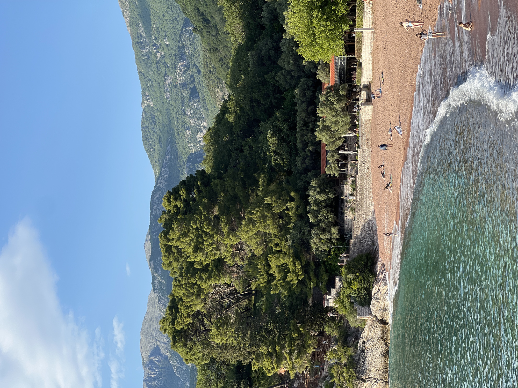 The image depicts a beach scene at Sveti Stefan, Montenegro. Azure water meets a sandy beach where several individuals are sunbathing or wading. A wave is crashing on the shore. Adjacent to the beach, a stone wall runs along the edge, lined with trees and structures that suggest the presence of restaurants or establishments. Behind the foliage rises a steep, green-covered mountain, contrasting with the clear, blue sky.