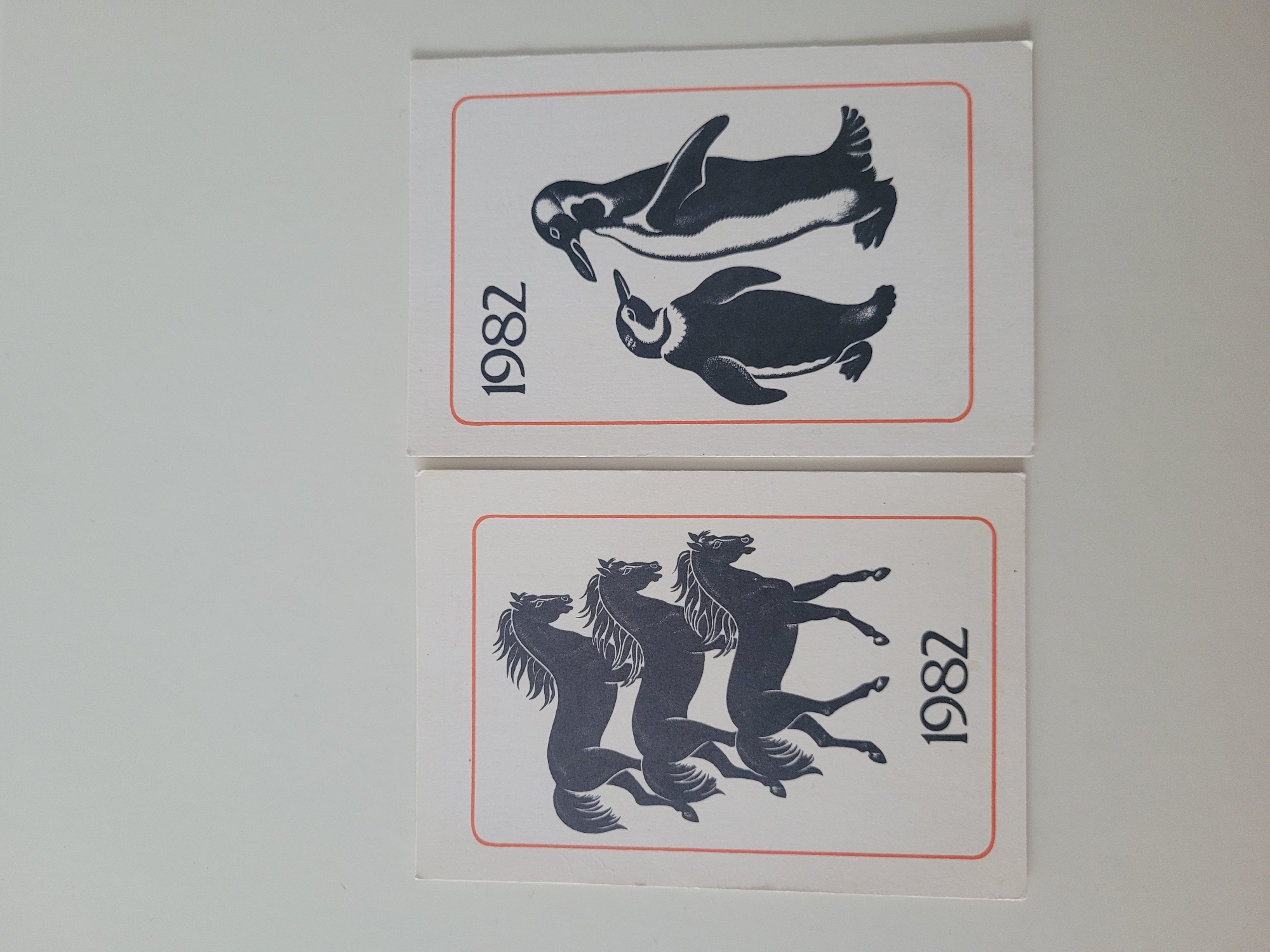 The image shows two cards against a white background. Both cards have a thin orange border. The card on the left features a dark grey illustration of three horses in profile, arranged vertically. Below the horses, the number "1982" is printed in dark grey. The card on the right features a dark grey illustration of two penguins, one larger and one smaller, standing side-by-side. Above the penguins, the number "1982" is printed in dark grey. The image is taken from a top-down perspective, with the cards lying flat. There is no indication of any specific location within the city of Jūrmala, Latvia.