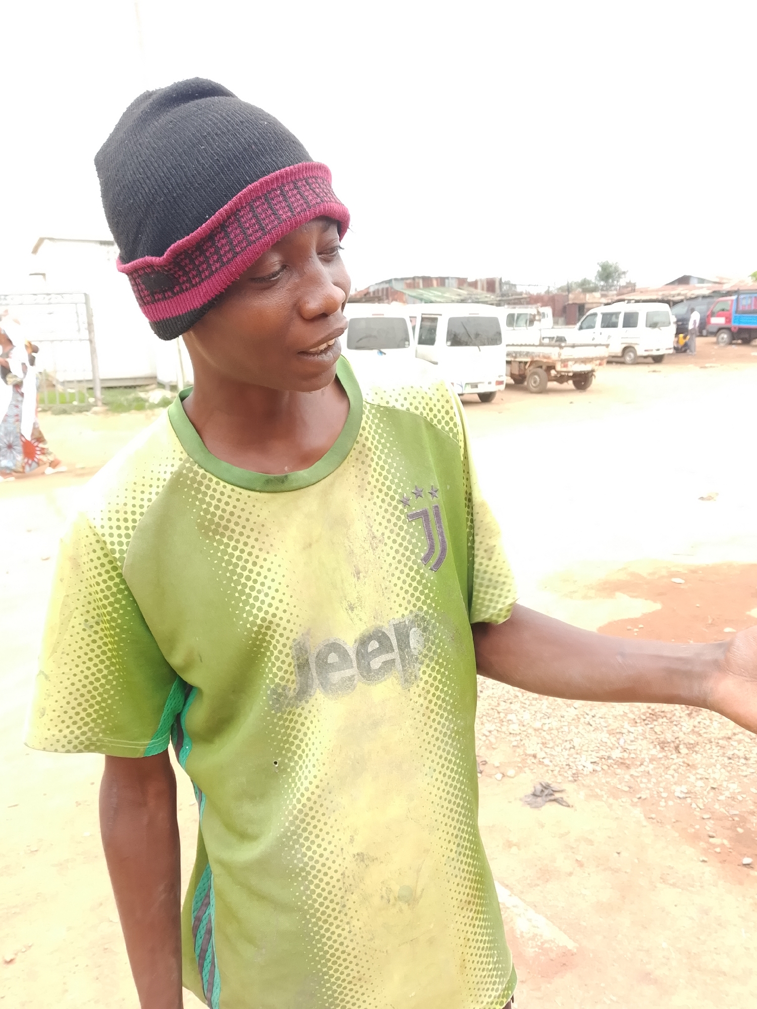 In an outdoor scene at a location in the city of Unknown, Unknown, a young person wearing a black beanie with a red and black patterned brim and a light green t-shirt with a pixelated pattern and the word "Jeep" emblazoned across the chest is pictured. The t-shirt also features a dark purple Juventus logo on the left chest. The person's head is turned to the right, and their mouth is slightly open, as if speaking or in mid-thought. Their right arm is extended outwards, with their hand open, palm facing upwards. In the background, several white vans and a small truck are parked on a dirt lot, suggesting a market or transportation hub. The sky is overcast, contributing to the muted lighting of the scene. A few other people are faintly visible in the distance.