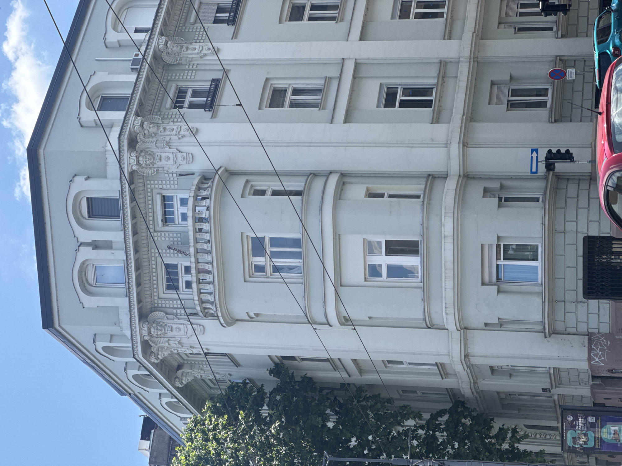 The image depicts a light blue, ornate building on a street corner in Belgrade, Serbia. The building features a curved facade with multiple windows of various sizes and styles, some with shutters. Decorative elements, including sculptures of faces and intricate moldings, adorn the upper levels of the building. Overhead wires crisscross the scene, indicating the presence of public transportation, likely trams. A large green tree partially obscures the left side of the building. Below, a traffic light with a rightward arrow sign is visible, alongside a red car in the foreground and a turquoise SUV parked behind it. A "no stopping" sign is also present. A poster advertisement for "Alma Quater" is displayed on the lower left. The sky is clear with a few wispy clouds.