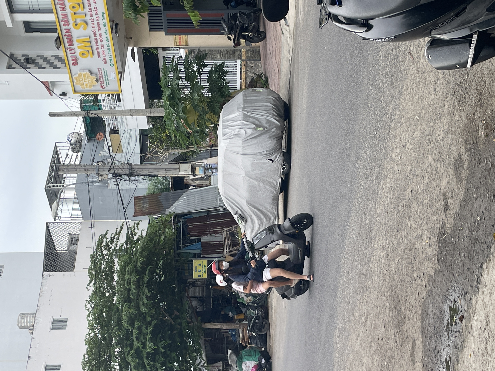 In Nha Trang, Vietnam, on a street with a cracked asphalt surface and a puddle of water on the left, a two-person scooter is traveling from left to right. The person in the front is wearing a red helmet and a white face mask, while the person in the back is wearing a white helmet and a white face mask. Both are wearing casual clothing. They are riding next to a car covered in a light grey tarp, which is parked on the right side of the road. Behind the car, a black motorcycle is parked on a sidewalk made of pinkish-red bricks. In the background, there are buildings with various architectural styles, including a white building with a water tank on the roof, a building with a gray facade, and a building with a sign for "OM STORE" in yellow lettering. There are also trees and power lines visible. The overall scene suggests a typical day in a Vietnamese city.