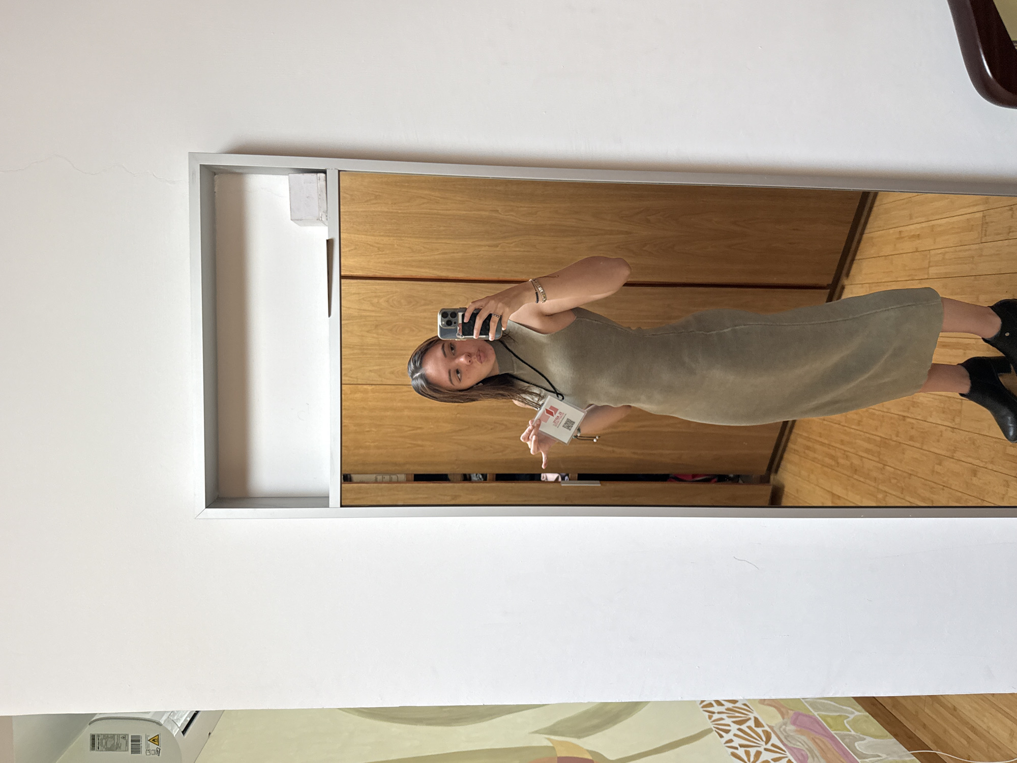 A woman is taking a selfie in a mirror. The mirror is framed in silver and reflects a scene of wooden cabinets and flooring. The woman is wearing a tan dress and black boots. She is holding a phone in her left hand and a badge in her right. The background outside the mirror shows a white wall with a crack and a colorful painting on the bottom. The location is San Bernardino Tlaxcalancingo, Mexico.