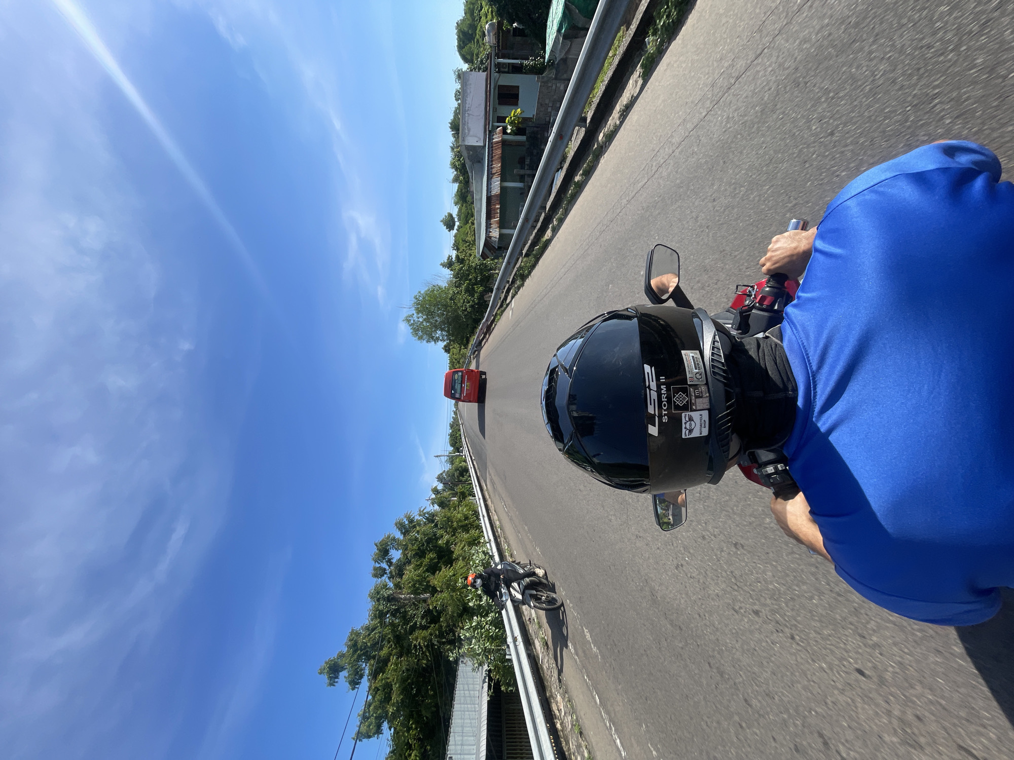 The image is a first-person perspective shot taken while riding a motorcycle on a road in Mui Ne, Vietnam. The rider is wearing a blue shirt and a black helmet. In front, another person on a motorcycle is visible, wearing a helmet and an orange face mask. Further ahead, a red bus is driving in the same direction. The road is asphalt, and there are metal guardrails on both sides. Lush green trees and some buildings are visible on the sides of the road, bordering the highway. The sky is blue with scattered white clouds. The overall scene depicts a sunny day on a road trip in a tropical location.