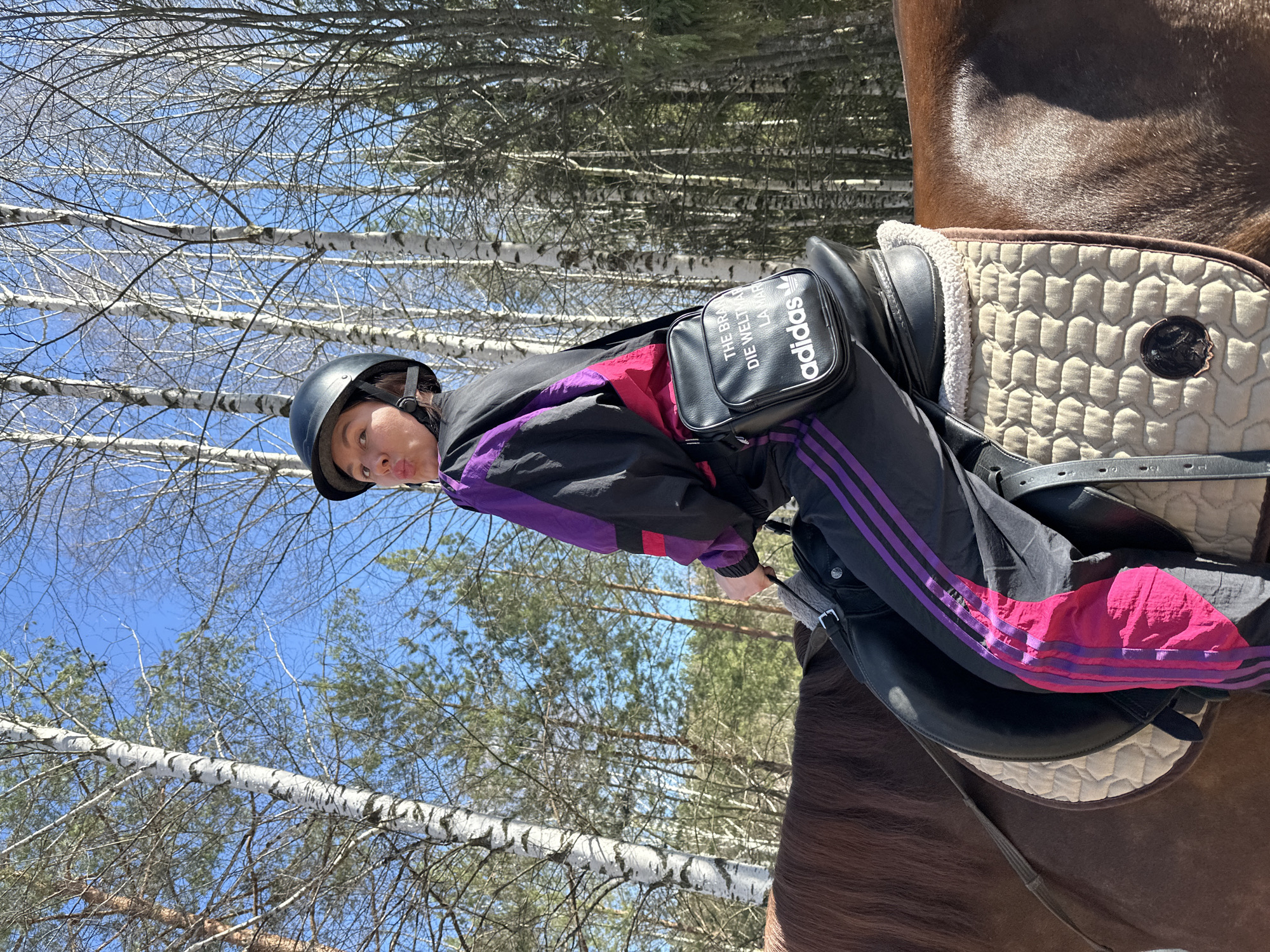 A person wearing a black helmet and an Adidas tracksuit is riding a brown horse. The horse is equipped with a saddle and a saddle pad. The scene is set in a forest with bare trees and a clear blue sky. The location is Русский Вожой, Russia.