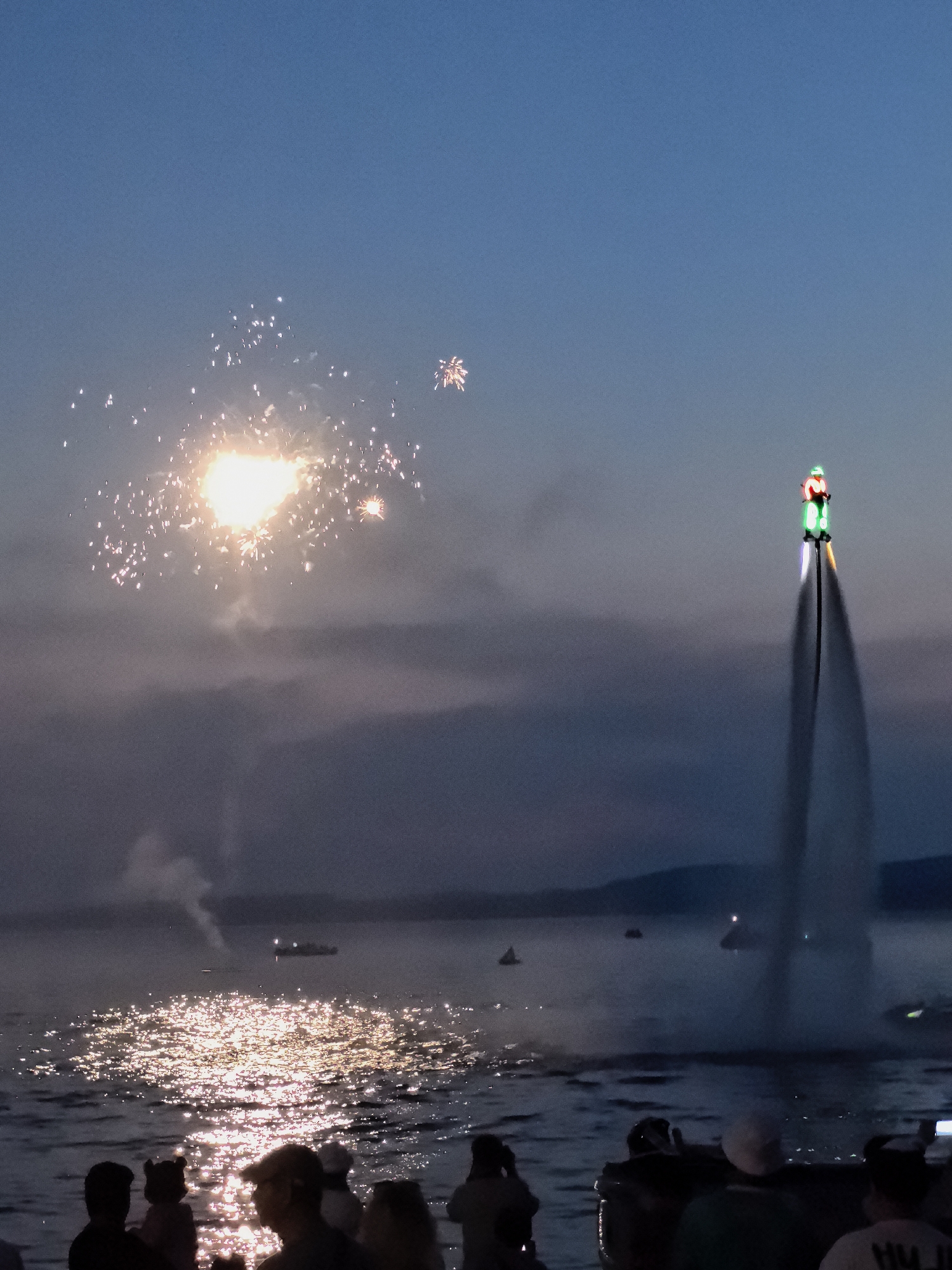 In Osa, Russia, during a nighttime event, a person on a jetpack, illuminated by green and red lights, ascends from the water. They are positioned on the right side of the frame, creating a prominent water stream beneath them. In the background, fireworks illuminate the sky with a bright burst of light on the left and smaller bursts of light scattered further into the distance. The calm water reflects the bright lights of the fireworks and a distant boat can be seen on the water. A crowd of people is gathered on the shore in the foreground, silhouetted against the water. The overall scene captures a celebratory atmosphere with elements of both pyrotechnics and aerial performance.