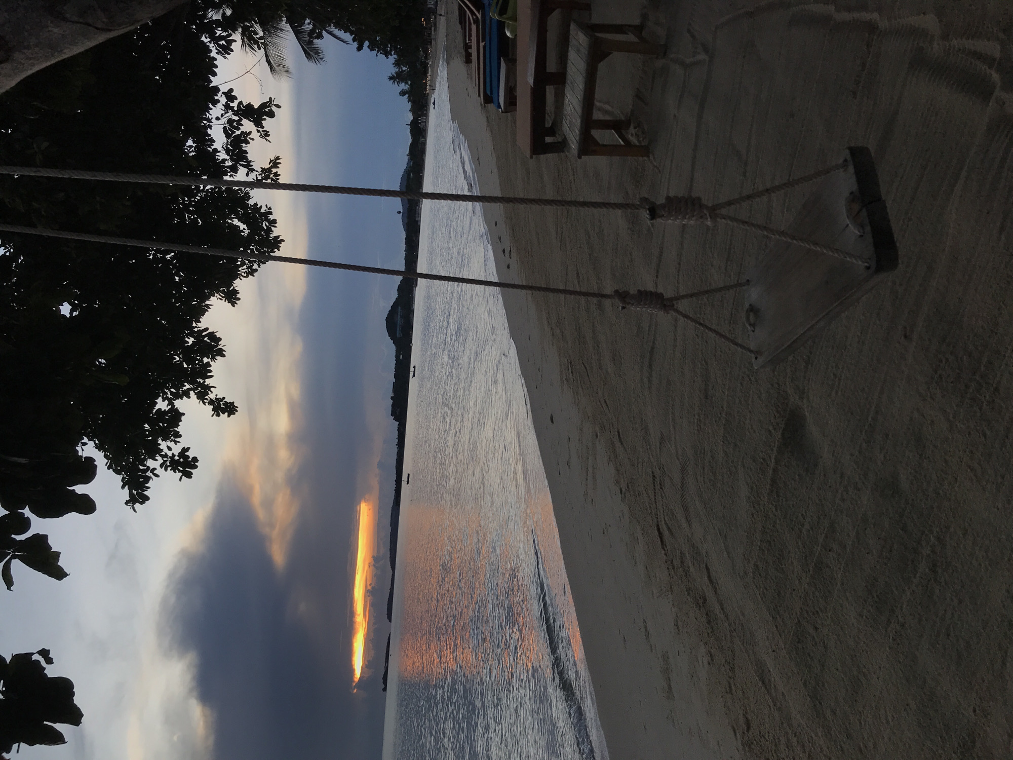 At a beach in Baan Mae Nam, Thailand, a rope swing hangs from a tree branch near the shoreline. A wooden bench sits on the sand near the swing. The sea reflects a sunset. The sky is partly cloudy.