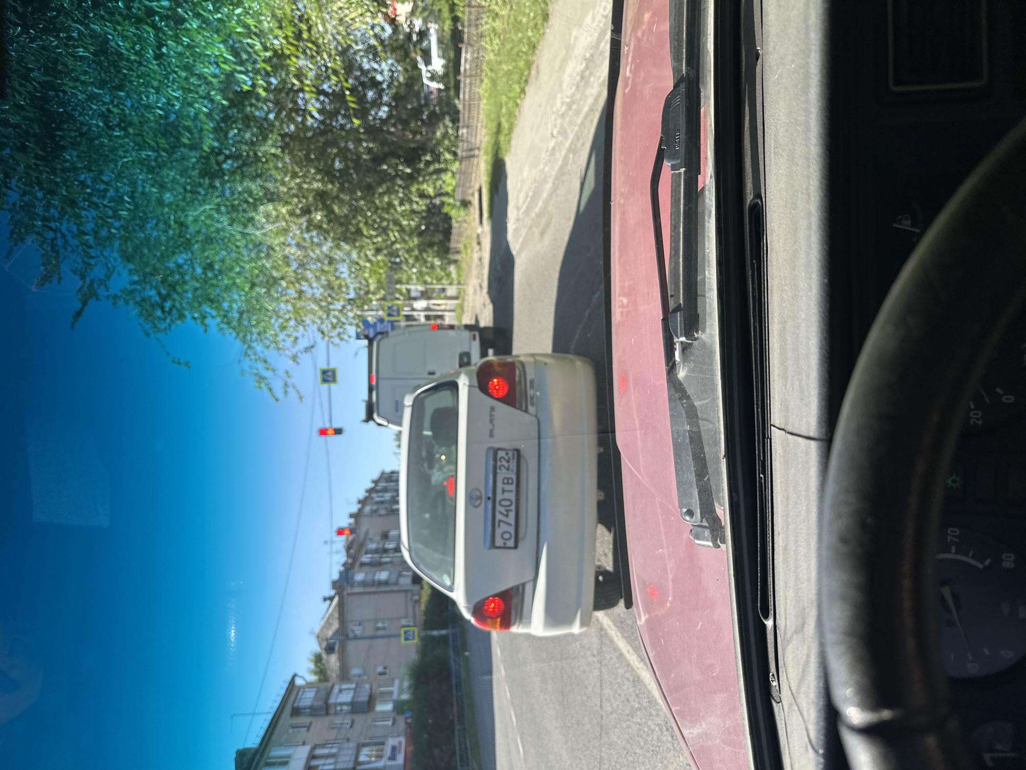 The image is a point-of-view shot from inside a vehicle, looking through the windshield at traffic on a street in Novoaltaysk, Russia. The camera is positioned behind a red car, as indicated by the car's hood and steering wheel visible in the foreground. In front of the camera is a white Toyota Platz sedan with a license plate reading "O 740 TB 22". Beyond the Toyota, a white cargo van is visible, followed by another vehicle. Buildings line the left side of the street, and a tree with lush green foliage is on the right. Traffic lights are red, indicating a halt. The sky is clear and blue. The environment appears to be a typical urban street scene in the summer.