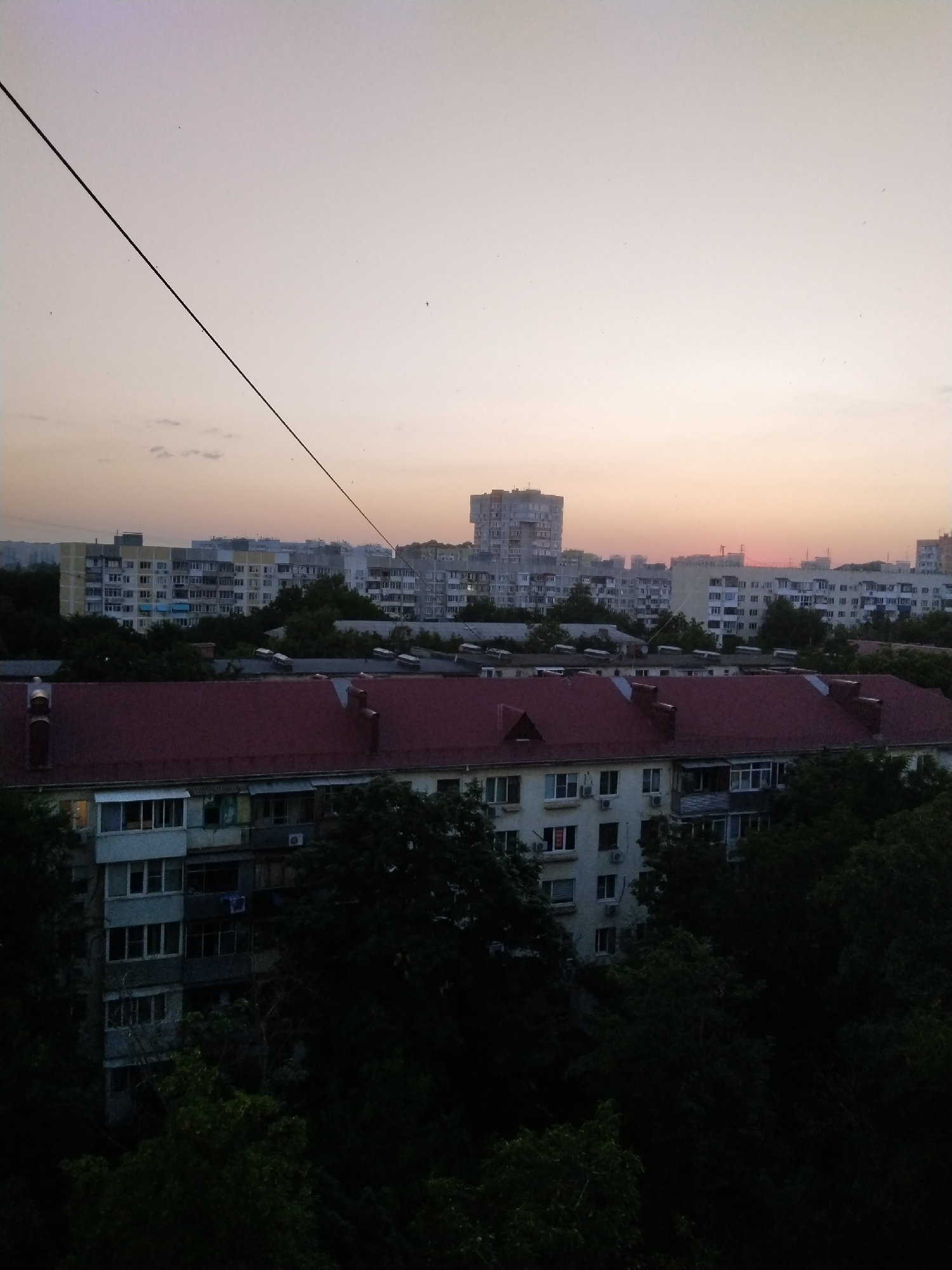 The image captures an urban landscape in Krasnodar, Russia, at dusk. The foreground is dominated by the dark silhouette of dense foliage, partially obscuring a multi-story apartment building with a prominent red-tiled roof. This building features numerous windows and balconies, some with visible air conditioning units and balconies enclosed with glass. Further in the background, a series of residential buildings of varying heights are visible. These buildings are characterized by their repetitive architectural style, common in Soviet-era urban planning, with rows of balconies and windows. A tall, distinct apartment block stands out among them. The sky above is a pale, gradient of pink and purple, suggesting the lingering light of sunset, with faint cloud formations and a few scattered birds visible. A thin, dark wire stretches diagonally across the upper portion of the frame.