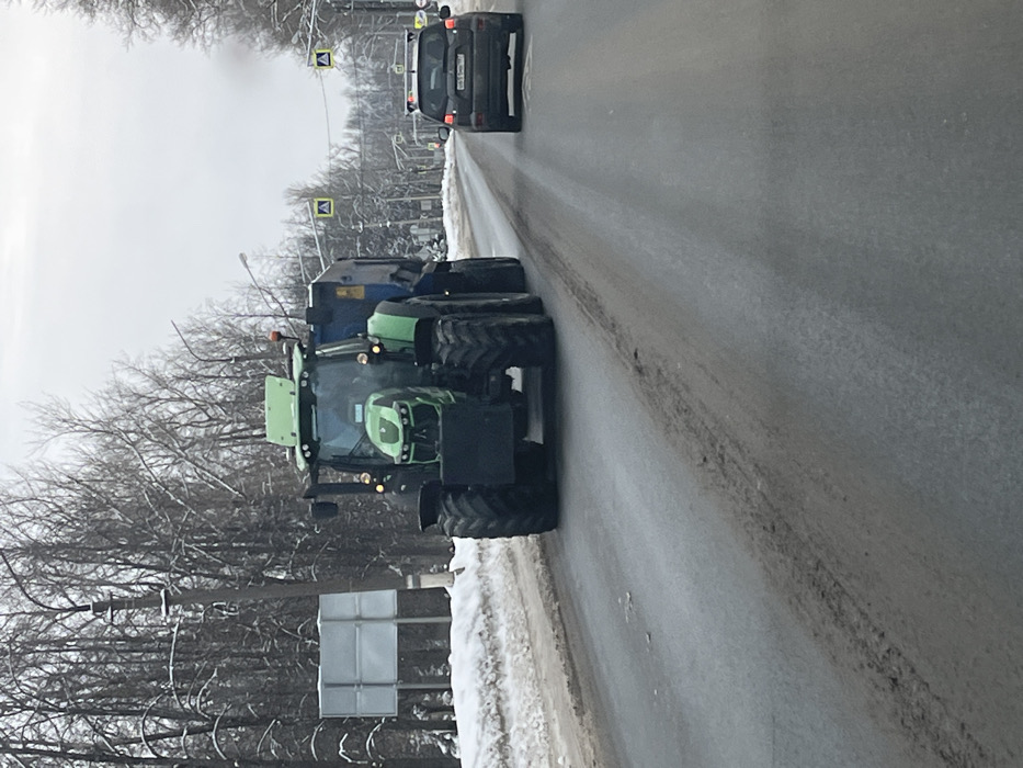 The image depicts a winter road scene in Novosinkovo, Russia. A large, green, modern agricultural tractor, identifiable as potentially a Deutz-Fahr model, equipped with a black front counterweight and towing a blue and black utility trailer, occupies the foreground and midground. The tractor's headlights and marker lights are illuminated, and a driver is discernible within the cab. The vehicle is traveling on a two-lane asphalt road that exhibits patches of dirt and slush, with accumulated snow banks lining the shoulders. Following the tractor in the same lane is a dark-colored SUV, displaying a Russian license plate and illuminated brake lights. The environment features bare deciduous trees and utility poles bordering the road. Several yellow diamond-shaped pedestrian crossing signs are suspended overhead across the roadway, and a large, blank rectangular sign stands on the left side of the road. The sky is uniformly overcast.