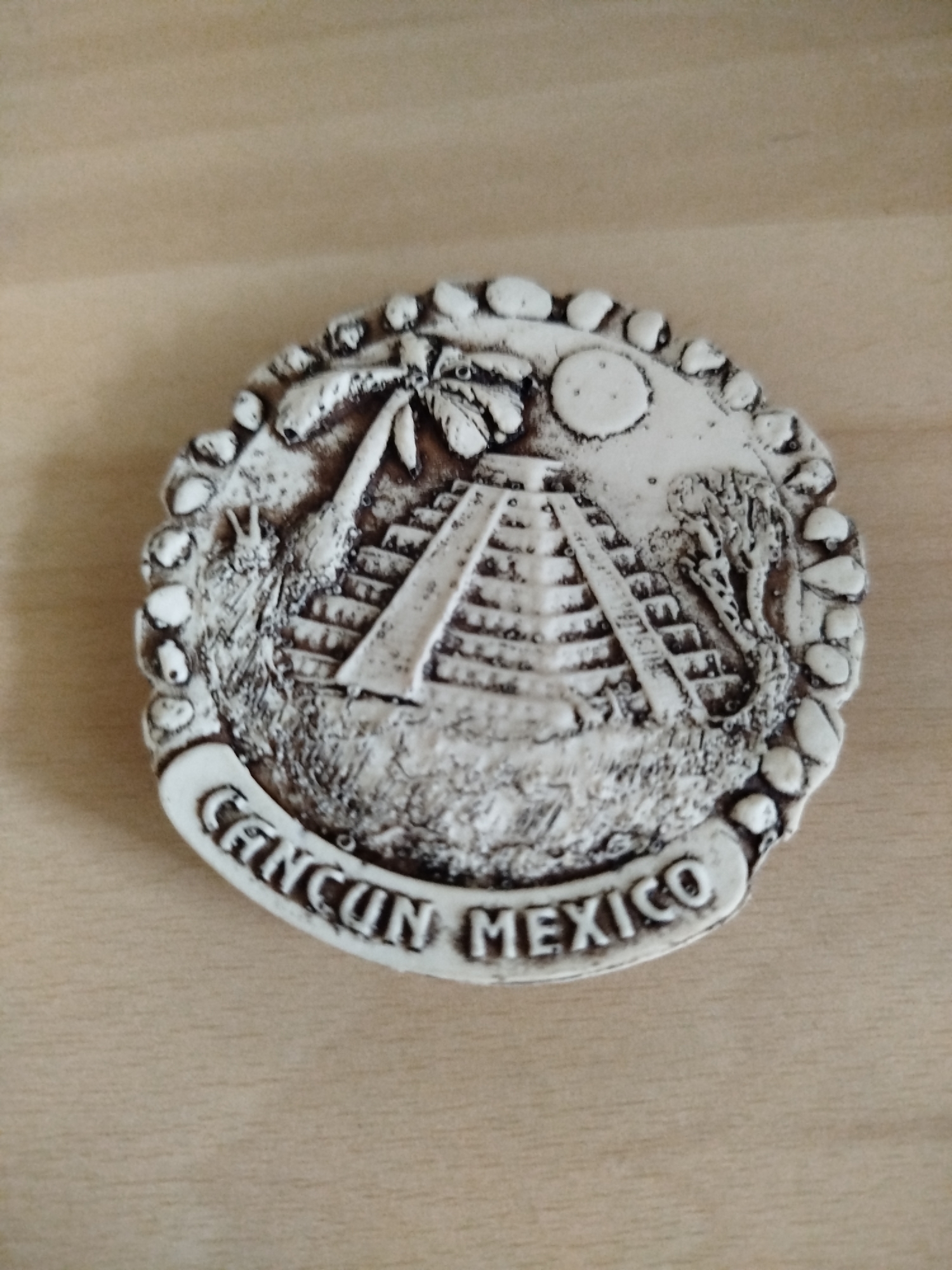 The image shows a souvenir magnet depicting a Mayan pyramid in Cancun, Mexico. The magnet is circular and off-white with a textured surface. It features a palm tree, a pyramid, a sun, and vegetation. The words "Cancun Mexico" are inscribed around the bottom edge. The background is a blurred, light brown, possibly wooden surface.