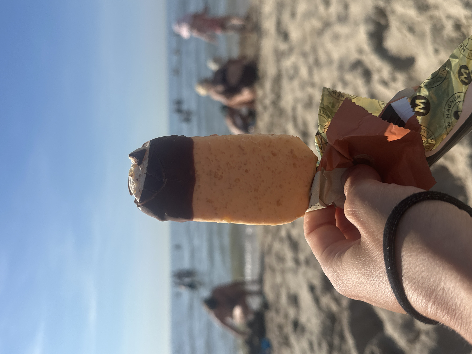 In Zandvoort, Netherlands, on a sunny day at the beach, a person's hand holds a partially unwrapped Magnum ice cream bar. The ice cream has a dark chocolate coating on top and a prominent orange-colored ice cream body. The background is blurred, showing the ocean with people swimming or wading in the water and others on the sandy beach. The sky is a clear, bright blue. The foreground focuses on the ice cream bar and the hand holding it, with the wrapper of the ice cream also visible.