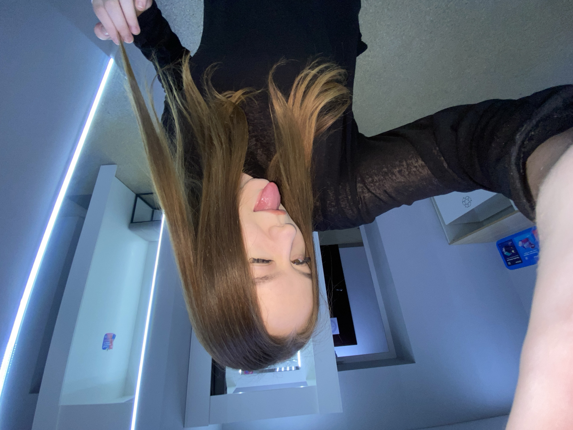 A young woman playfully poses for a photo in a modern, well-lit indoor setting, located in Chaikovsky, Russia. She is depicted from the chest up, with her head tilted, revealing her long, light brown hair cascading around her face and shoulders. She wears a black, long-sleeved top that appears somewhat sheer. Her expression is mischievous, with her tongue sticking out and her right eye winking. Her left hand gently holds a strand of her hair.
The background features a clean, contemporary design with prominent vertical LED light strips illuminating white walls or shelving units with a cool blue-white glow. To the right of her, a small blue object or screen with indistinct graphics is visible. Near the bottom center of the frame, on a white surface, a black recycling symbol (three arrows forming a triangle) is clearly visible. The artificial lighting and sleek decor suggest a modern room, possibly a home, studio, or office, creating a vibrant and cheerful atmosphere for the playful pose.