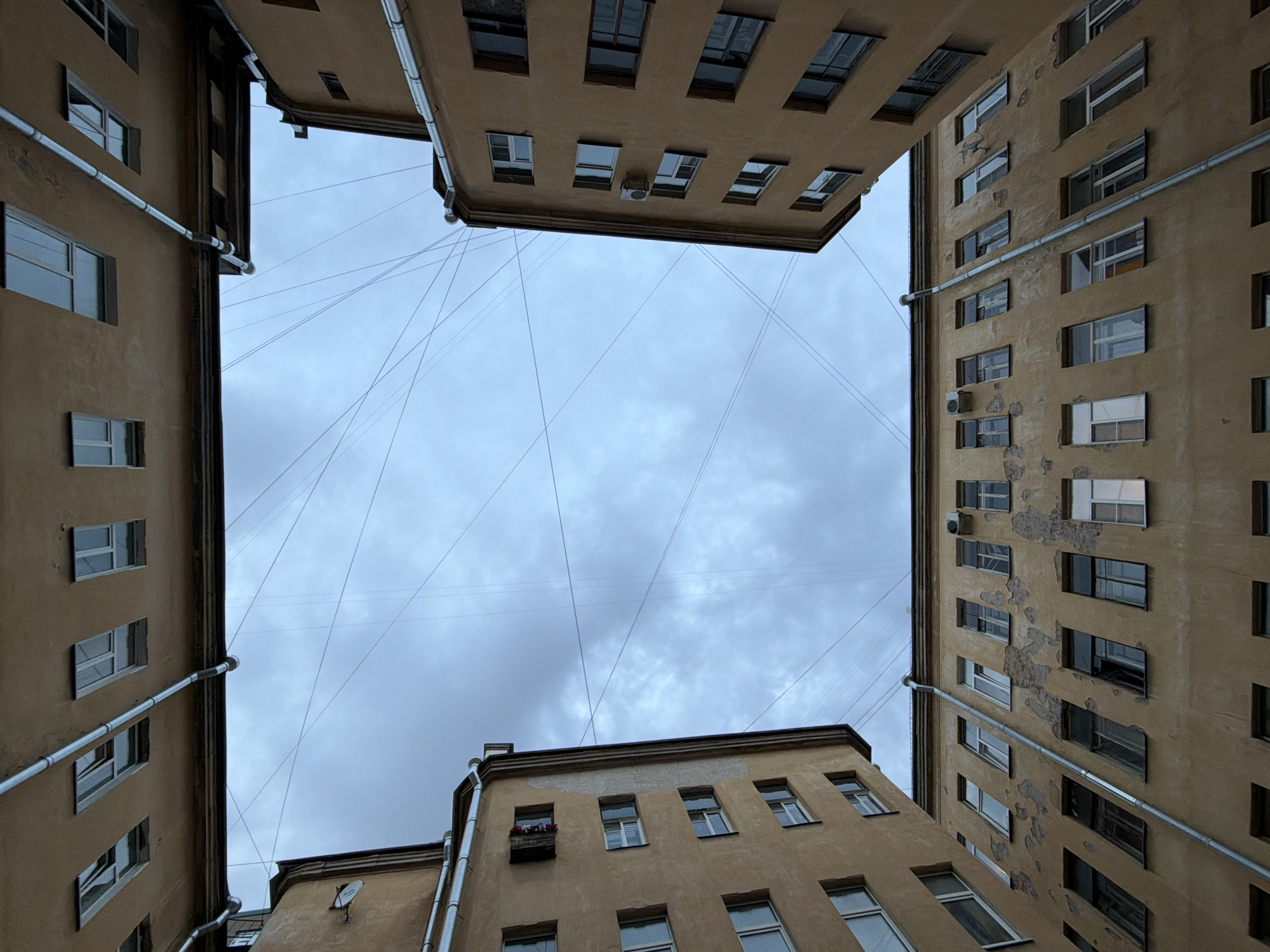 This is an upward-facing shot of a courtyard in Saint Petersburg, Russia, characterized by its "well" or "passage" architecture. The buildings on all four sides of the courtyard are visible, appearing aged with peeling paint and a uniform beige color. Multiple rows of windows are present on each building facade. Overhead, a cloudy sky is crisscrossed by a network of wires. Gutters and downspouts run along the exterior walls of the buildings. The perspective suggests the image was taken from the ground level within the courtyard, looking directly upwards. There are no people visible in the frame.