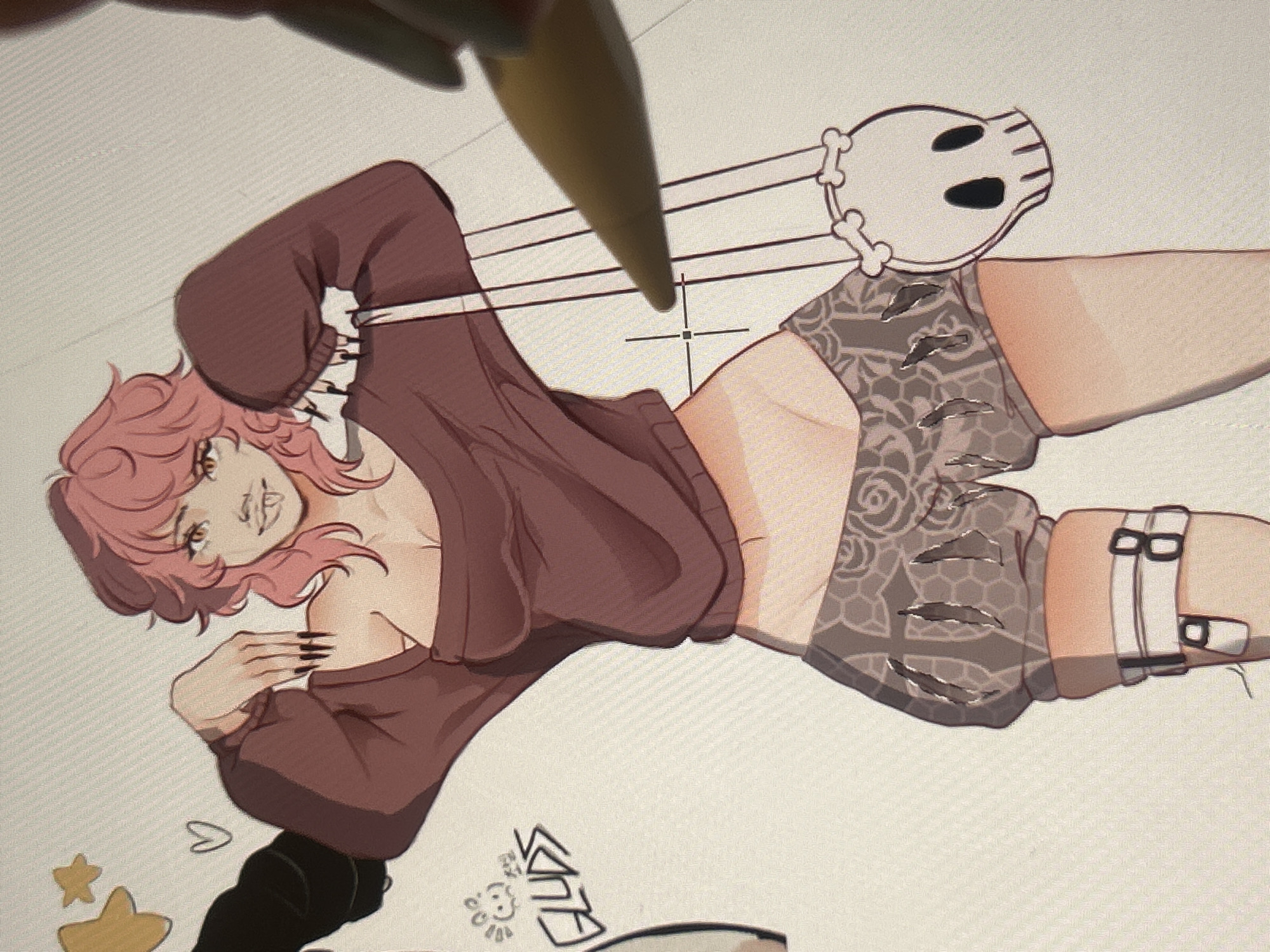 This is a digital artwork in progress, displayed on a screen. The main subject is a stylized anime character with pink hair, wearing a loose-fitting maroon sweater and patterned shorts. The character is depicted in a dynamic pose, with one arm bent and hand near their face, and the other arm extended. A prominent detail is a skull accessory with bone-like straps attached to the character's thigh. The artwork also features several stars and a heart drawn in a simpler style on the lower left. Some text, stylized and illegible, is visible in the lower right and center of the image. A stylus is actively being used to draw or edit the artwork, casting a shadow. The overall aesthetic suggests a fantasy or edgy theme. The digital interface grid is visible in the background, indicating it is a work in progress on a digital canvas. There are no overt indications of time of day, weather, or specific location cues beyond the general context of digital art creation. The character's expression is ambiguous, but the overall mood is somewhat alluring.