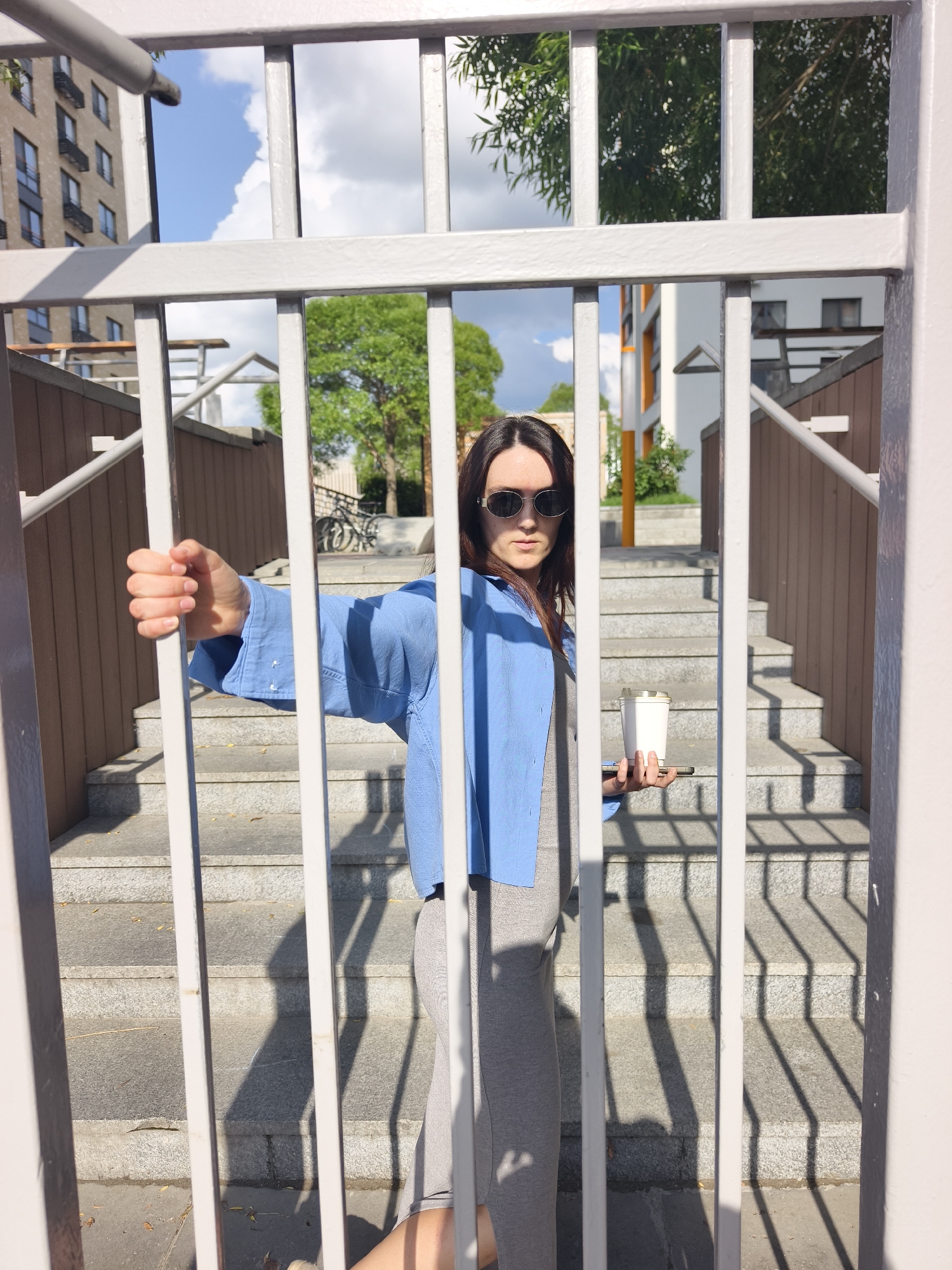 The image captures an outdoor urban scene in Tyumen, Russia, under bright daylight conditions, evidenced by prominent shadows. A single adult female stands in the immediate foreground, positioned behind a series of six evenly spaced, vertical light-colored metal bars, which are part of a fence or gate. She has long, dark hair and wears dark, oval-shaped sunglasses. Her attire includes a light blue, long-sleeved collared shirt or jacket over a gray, form-fitting sleeveless dress or top. Her left hand is closed around one of the vertical bars. In her right hand, she holds a white, lidded cup, which rests on a horizontally oriented smartphone.
The background consists of a multi-level outdoor staircase constructed from light gray concrete or stone, featuring both brown and white railings. To the far left, a multi-story residential building with brown or beige cladding and dark windows is visible. Towards the center-right, a contemporary white building with orange architectural details can be seen, partially obscured by green deciduous trees. A bicycle is faintly discernible near the upper portion of the stairs in the mid-ground. Strong, elongated shadows from the foreground bars are cast across the concrete ground and steps. The sky above is partially visible, displaying clear blue patches interspersed with white cumulus clouds.