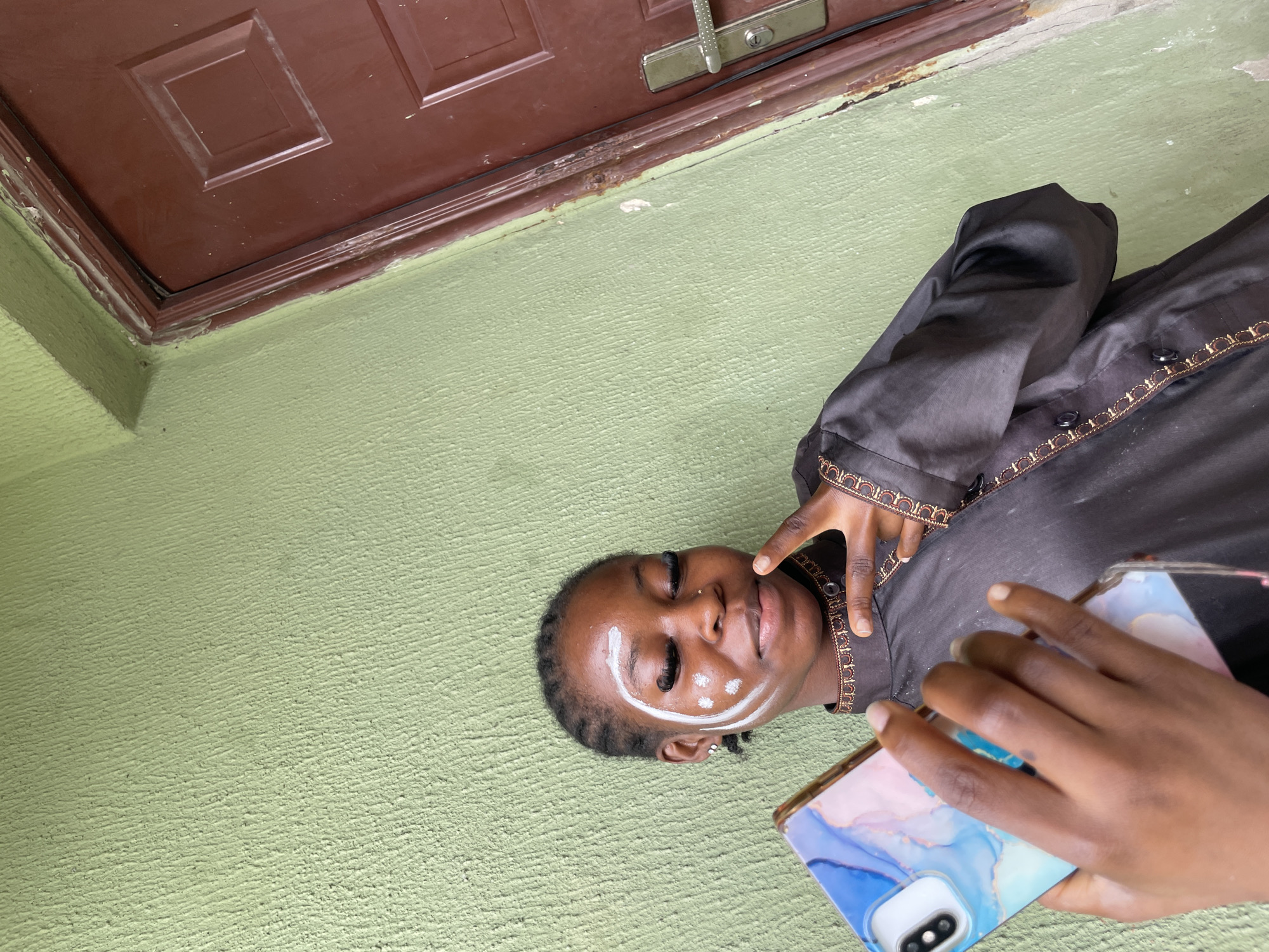 A young woman with braided hair and white face paint is holding a smartphone in her left hand. She is wearing a dark, collared shirt with gold trim. The woman is standing in front of a textured, light green wall and a brown door. Her right hand is held up in a peace sign. The scene appears to be outdoors, possibly in Ado, Nigeria.