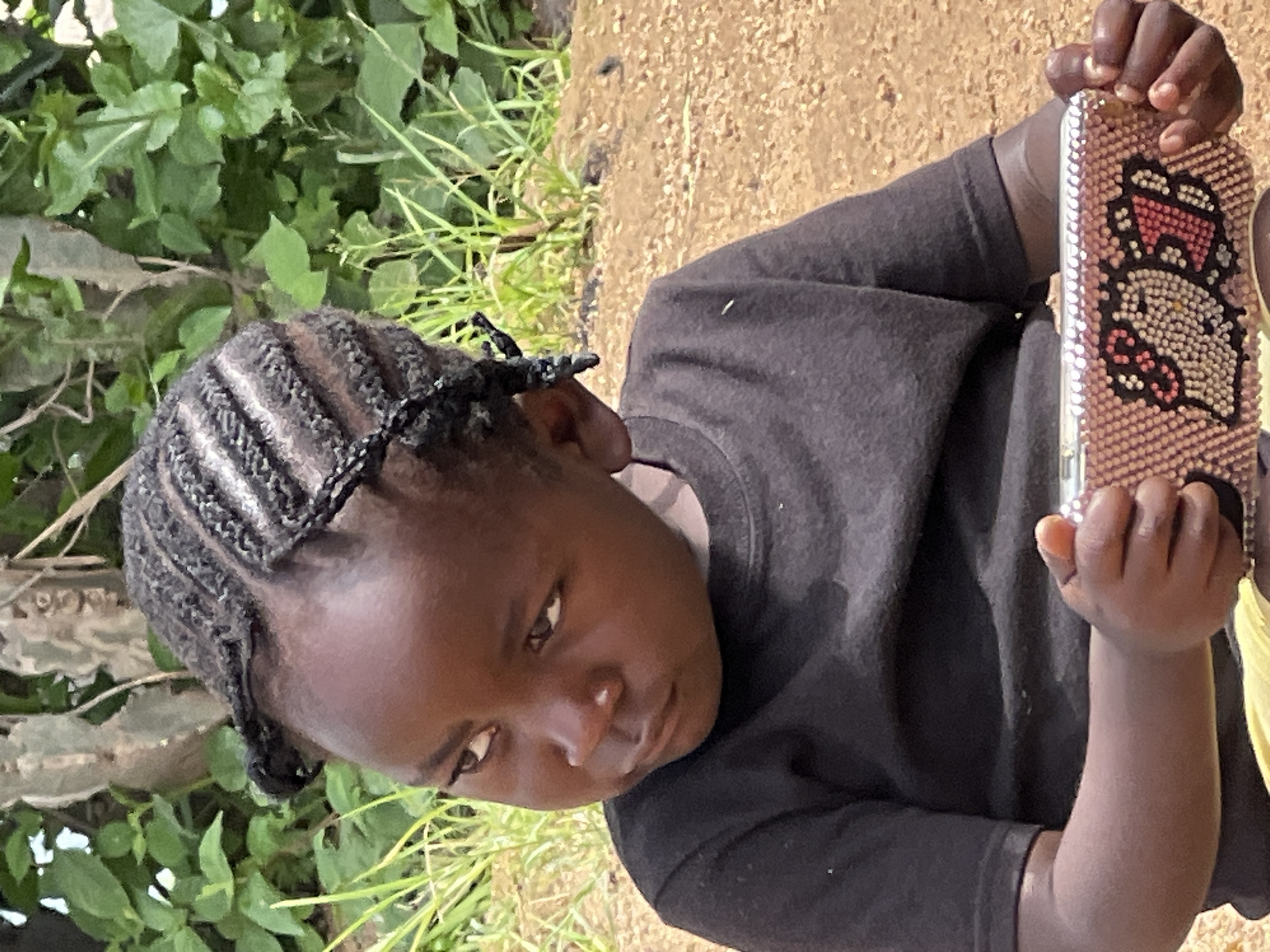 A young child with dark skin and braided hair is shown in a medium shot. The child is wearing a black t-shirt and yellow shorts. They are holding a pink phone case decorated with a cartoon character. The background consists of green foliage and brown dirt. The scene appears to be outdoors in a natural or semi-rural setting, possibly in Jos, Nigeria. The child is looking to their left, with a thoughtful expression. The lighting suggests it is daytime.