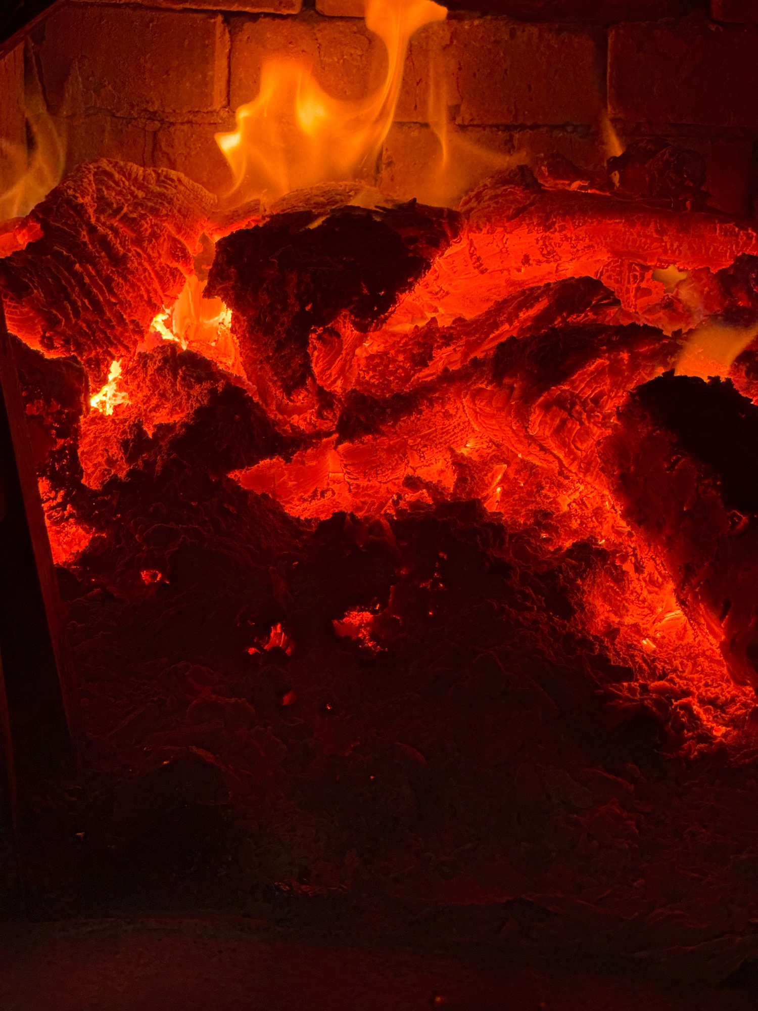 The image shows a close-up, low-angle view of a fireplace filled with glowing embers and flames. The wood is burning intensely, with vibrant orange and red hues dominating the scene. The brickwork of the fireplace is visible in the background, also illuminated by the firelight. The composition is abstract and focuses on the textures and colors of the burning wood. There is no indication of the city of Rusalevka, Russia, or any people in the image.