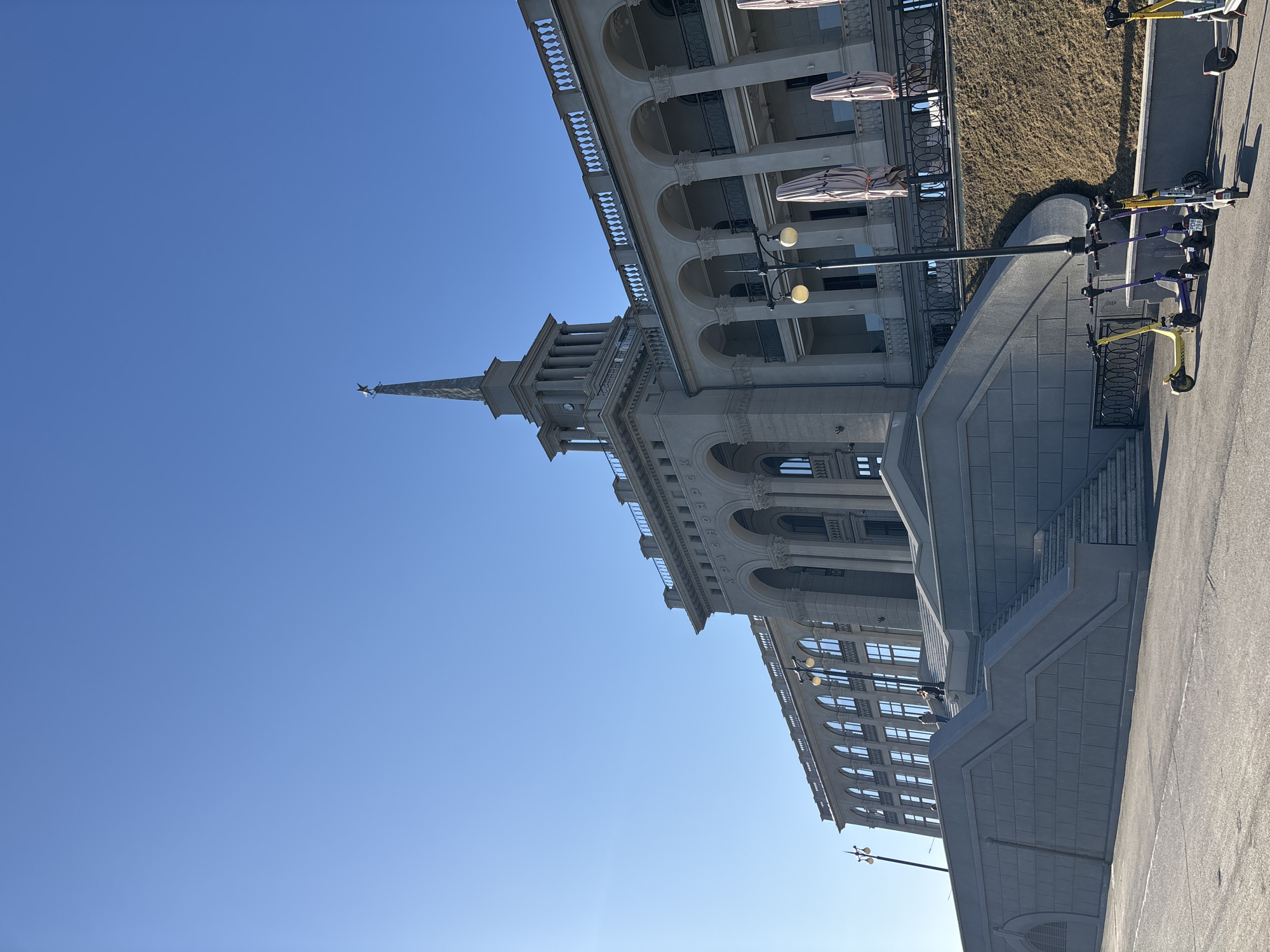 The image shows a building in Krasnoyarsk, Russia. The building has a tall spire with a star at the top. The building is made of gray stone and has many windows and arches. There are stairs leading up to the building. Several electric scooters are parked on the right side of the image. The sky is blue and clear.