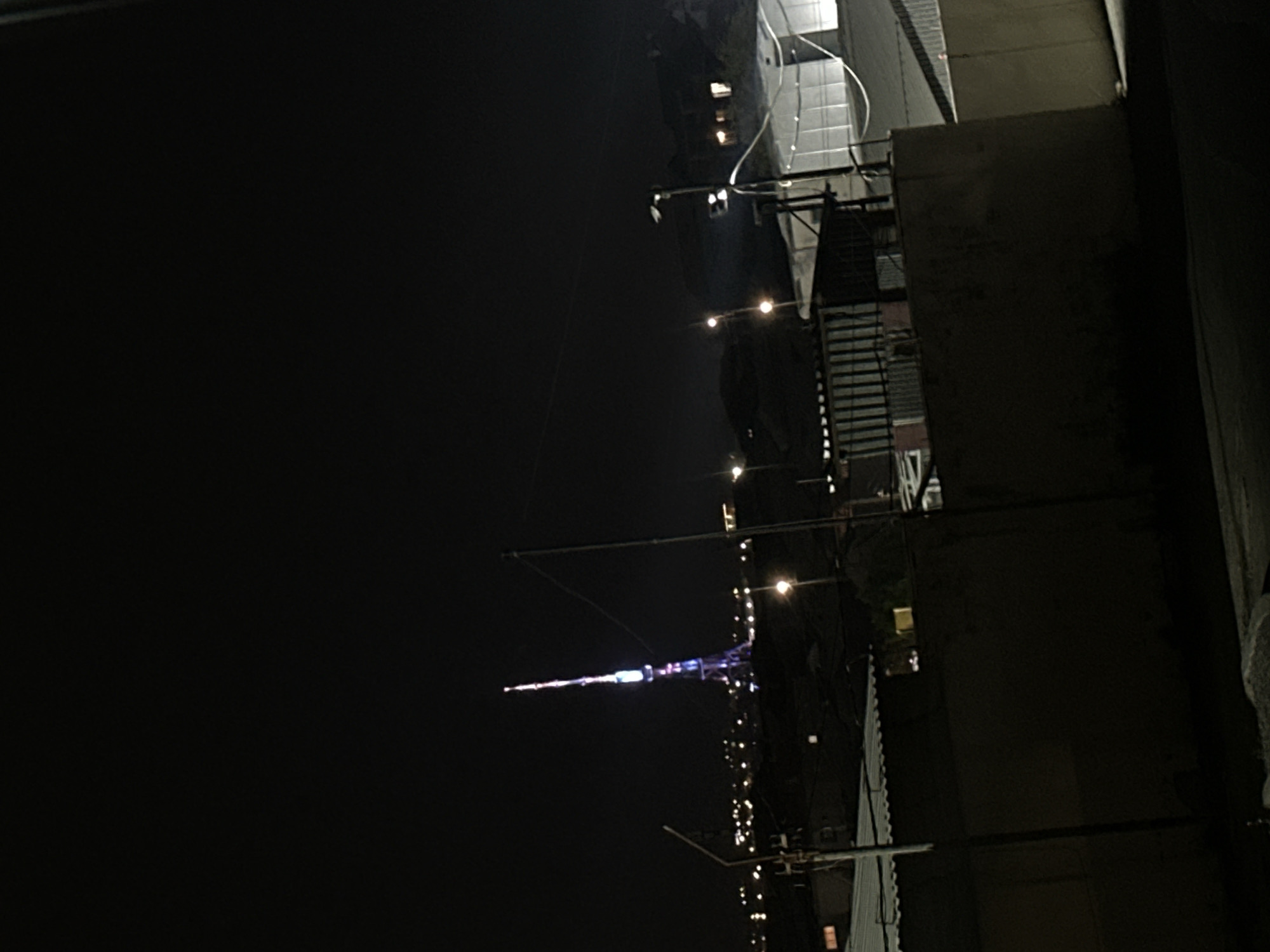 The image captures a nighttime urban or suburban scene, viewed from an elevated perspective. The central subject is a tall, slender tower, prominently illuminated in various colors including purple, blue, and white. This tower stands out against the dark night sky, dominating the mid-ground of the composition.
To the right and in the foreground, parts of several structures, likely building rooftops or balconies, are visible. These structures appear dark but are adorned with numerous small, warm-toned lights, possibly string lights or individual fixtures, creating a festive or atmospheric glow. Wires or cables are discernible running across the frame, connecting elements of the foreground architecture. The time of day is clearly night, with the sky appearing completely dark and cloudless. There are no people visible, and no specific activity is taking place. No visible text is present in the image.