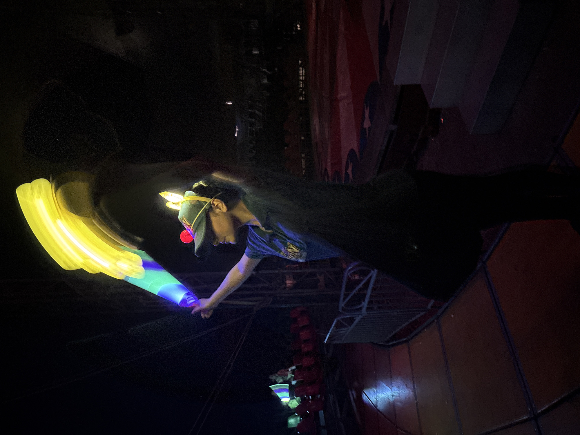A person wearing a cap with a red button is shown holding a blurred yellow and blue light stick. The person is wearing a dark-colored t-shirt and appears to be standing on a metal platform. In the background, a wall with red, white, and blue paint is visible, along with the structure of what looks like a stage or building. Hanging lights are also visible in the background. Based on the metadata, the image was taken in Tejalpa, Mexico.