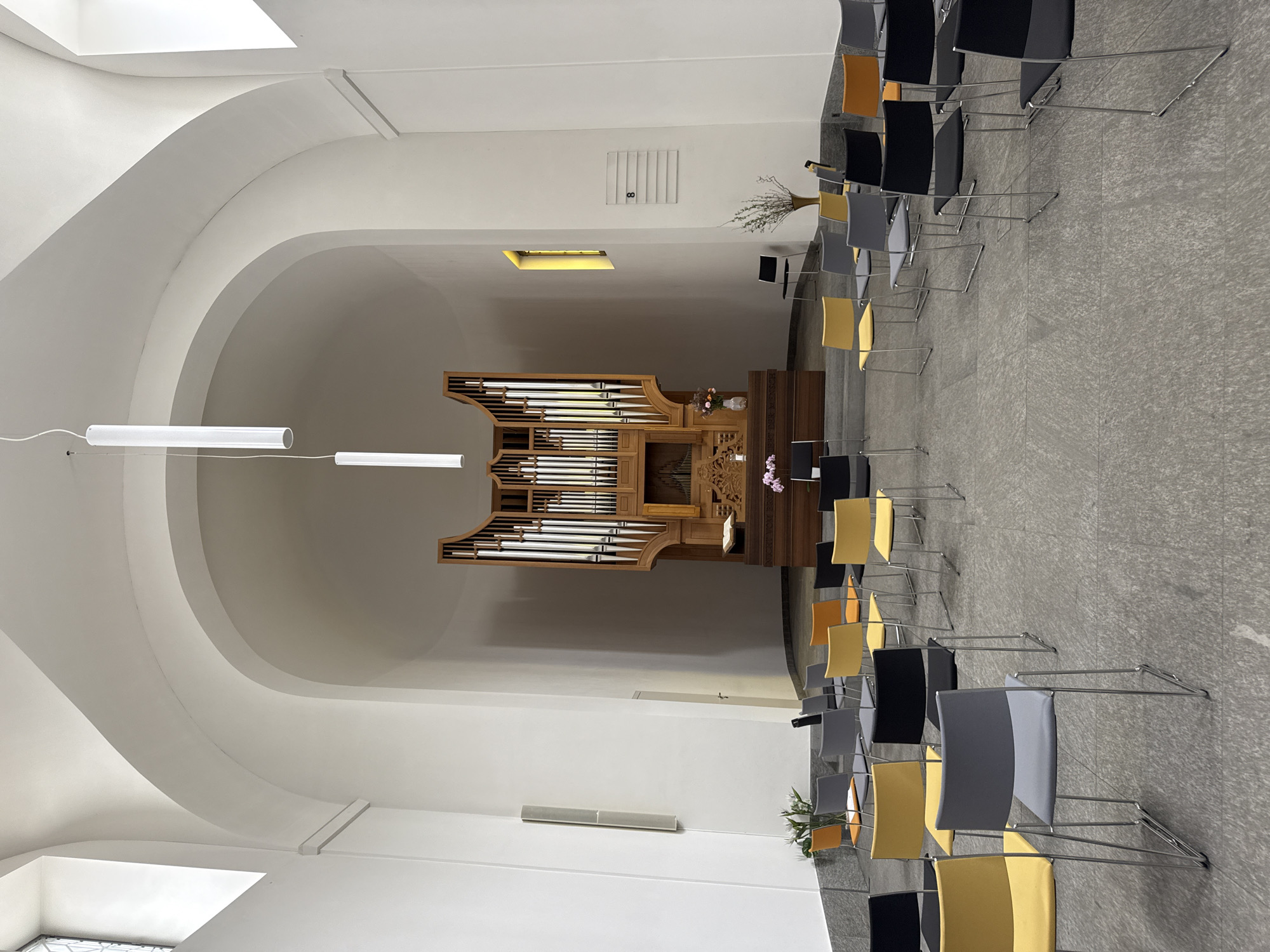 The image shows the interior of a church in Sesto San Giovanni, Italy. The main focal point is a large wooden organ with intricate carvings situated at the far end of the room, beneath a curved ceiling. Rows of chairs in various colors, including yellow, grey, and black, are arranged on the tiled floor facing the organ. Some chairs are empty, while others appear to be occupied by the back of unseen individuals. In front of the organ, there is a wooden altar with a book placed upon it, and floral arrangements. The walls are white, and the room is lit by overhead fluorescent lights and a small, yellow-tinted window on the right. The architecture features arched ceilings and large windows on the left. A sign with the number "8" is mounted on the right wall. The overall atmosphere is one of quiet reverence and a minimalist aesthetic.