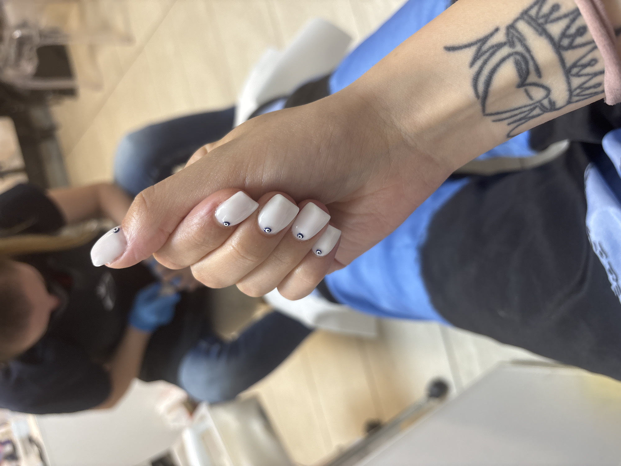 A woman's hand with white, square-shaped acrylic nails is featured prominently in the center of the frame, each adorned with a single, small black and white design. The woman has a tattoo on her wrist. The woman is seated, with blue and black clothing visible. A second person, possibly a nail technician, wearing a blue glove is visible in the background to the left, focusing on the woman's hand. The scene appears to be in a nail salon. The location cannot be determined from the information available.