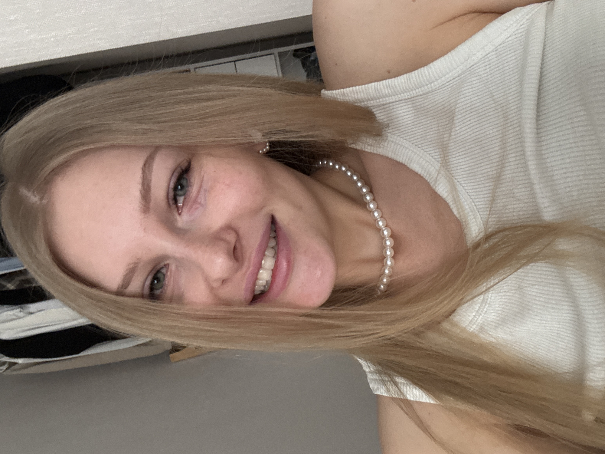 A young, light-skinned woman with blonde hair is smiling at the camera. She is wearing a white ribbed tank top and a pearl necklace. The background includes a closet or shelf with visible clothing and neutral-toned walls. The location is not discernible from the image.