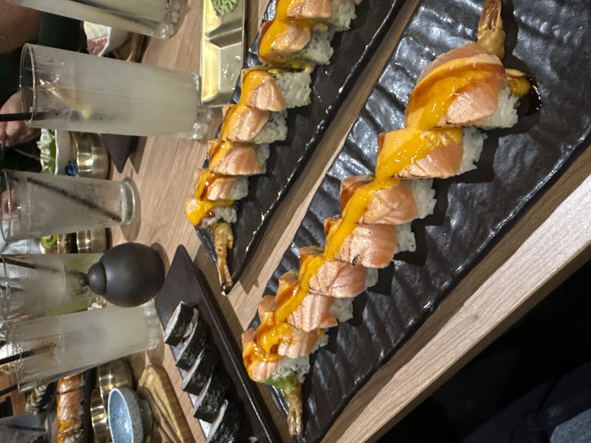 The image depicts a restaurant table setting with sushi and drinks. The sushi is arranged on two rectangular black plates, featuring cooked salmon slices drizzled with yellow sauce. The sushi rolls include rice, avocado, and fried shrimp tails. There are multiple glasses of what appears to be lemonade with straws. Additional sushi rolls are on a smaller plate, and there are also a black container, small dishes, and other tableware visible. The table has a light wood grain finish. The location is possibly a restaurant in Southlake, United States of America, based on the context of the task request. No people are directly visible, but the presence of drinks and partially eaten food indicates they are dining. The scene is indoors, suggesting a restaurant environment.