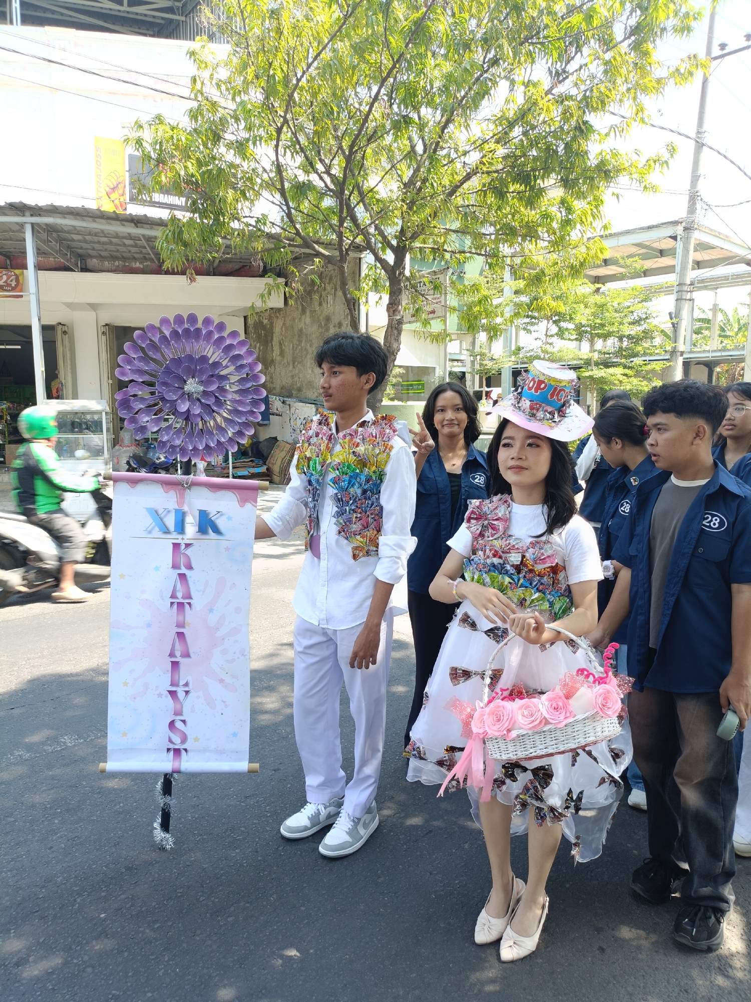 In Kediri City, Indonesia, a group of young people are participating in a parade. Two individuals in the foreground are dressed in elaborate costumes. The male on the left is wearing a white shirt and pants, adorned with a colorful vest made of what appears to be candy wrappers. He is holding a banner that reads "XIX KALYST". To his right, a female is wearing a white dress and a hat decorated with a soda can. Her dress is also embellished with candy wrappers. She is carrying a white basket filled with pink roses. Behind them, other participants in blue shirts with the number "28" are visible. In the background, there are buildings, trees, and a motorcycle with a rider wearing a green helmet. The scene suggests a festive event or cultural celebration.