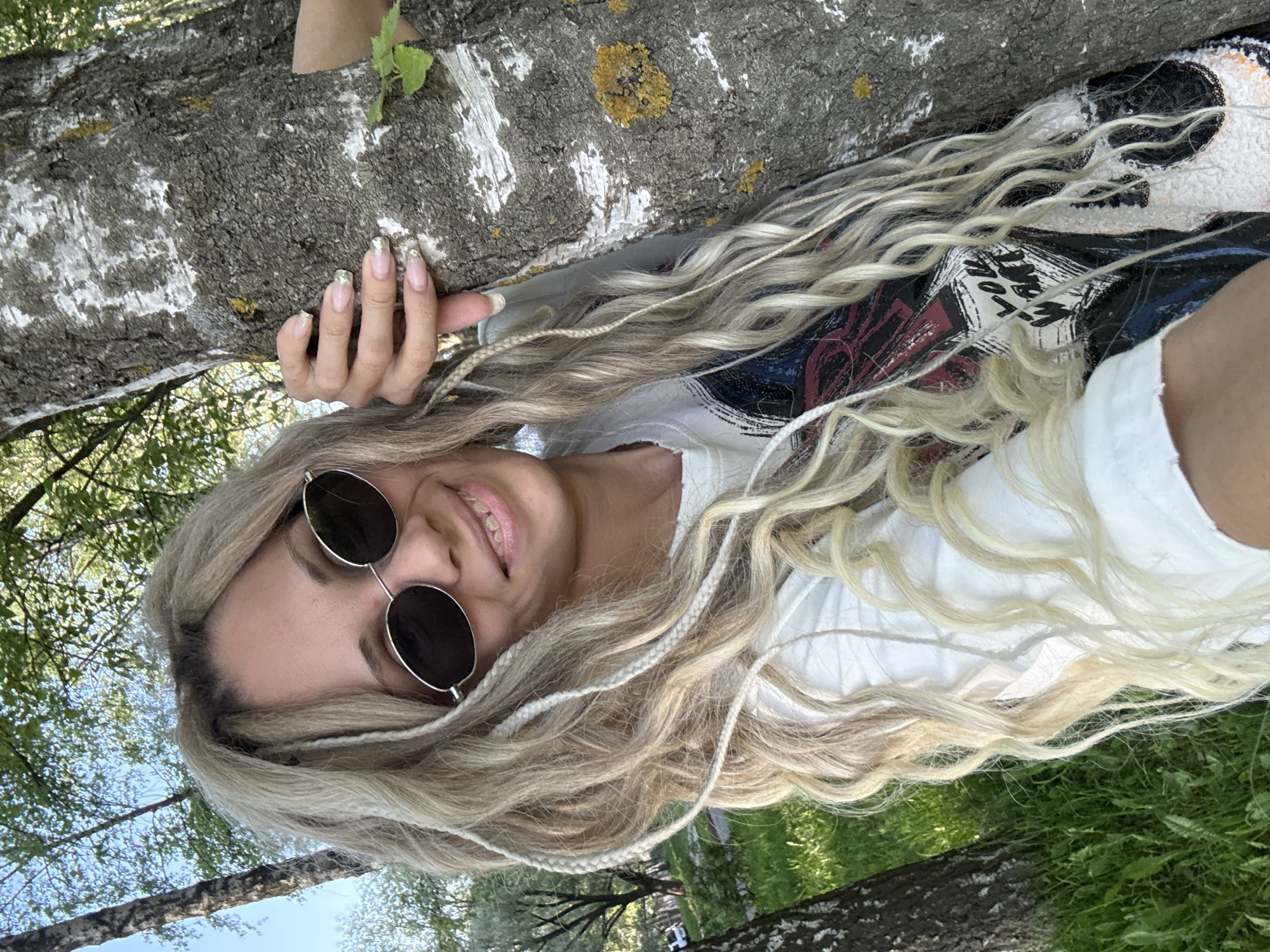 A fair-skinned female poses in a rotated selfie in what appears to be a park. The subject is wearing round sunglasses, a white shirt, and has long blonde hair with several small braids. She is smiling slightly. Her left hand is gripping a tree trunk with mottled gray bark. The tree trunk is also covered in patches of yellow lichen. Patches of green grass are visible in the background. Based on the angle and positioning, it appears the female is lying down. It is daytime. The location is Rusanovo, Russia.