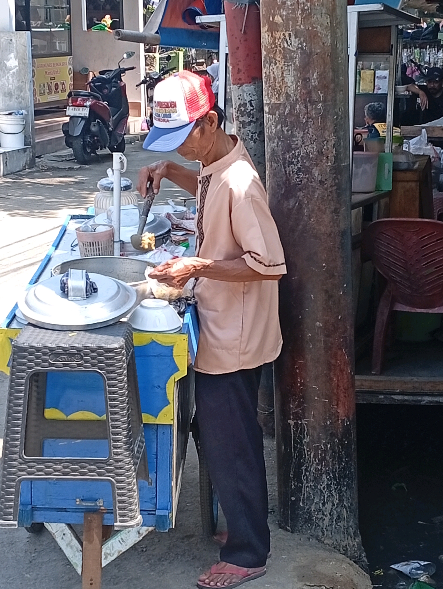 An elderly Indonesian man wearing a light peach-colored shirt and dark pants stands beside a food cart. He is wearing a red, white, and blue baseball cap with text that appears to read "PRESIDEN" and "BEM." In his right hand, he holds a ladle, which he is using to scoop food from a metal container on the cart. He appears to be in the process of serving or preparing food.
The food cart itself is predominantly blue with yellow accents. A black plastic stool is placed in front of the cart. Behind the man, a large, rough-textured pole stands prominently. In the background, a motorcycle is parked. Further back, a building with a sign that includes Indonesian text is visible. To the right of the pole, a dark opening or space is present, and further to the right, a red plastic chair is visible. The ground appears to be paved. The overall scene suggests a street-side food vendor operation, likely in a public area.
The presence of a motorcycle with an Indonesian license plate, along with the architectural style and the clothing of the people, strongly suggests the location is in Indonesia. Based on the distinctive red and white Indonesian flag colors often seen on license plates and the general urban streetscape, and considering the prompt specifically mentions East Jakarta, Indonesia, this scene is consistent with a location within that region.