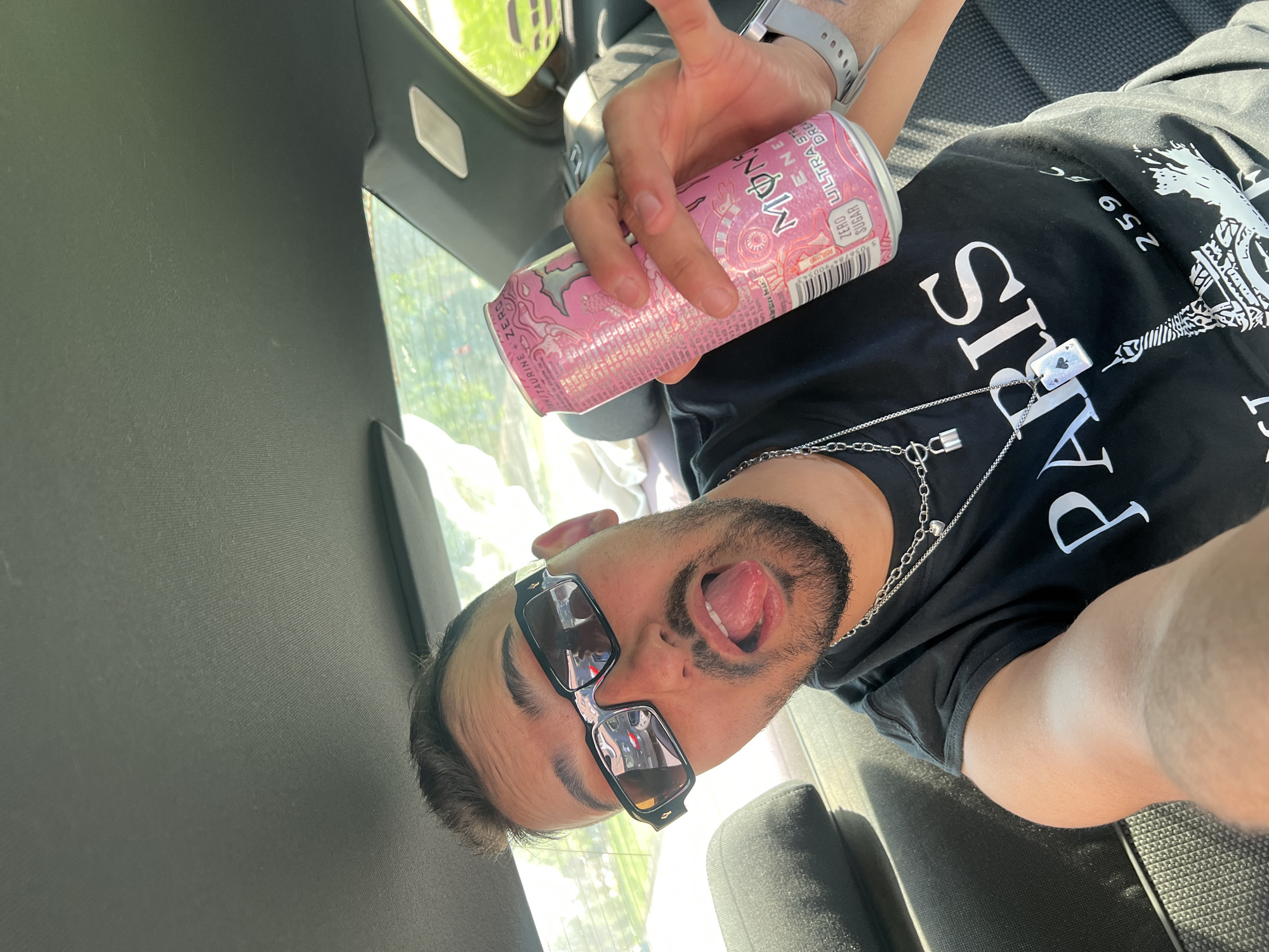 The photograph, taken inside a car, features a man wearing sunglasses and a black T-shirt with the word "PARIS" emblazoned across the front. He is holding a pink Monster energy drink can. His tongue is sticking out, and he is gesturing with his left hand. The location is Sevlievo, Bulgaria.