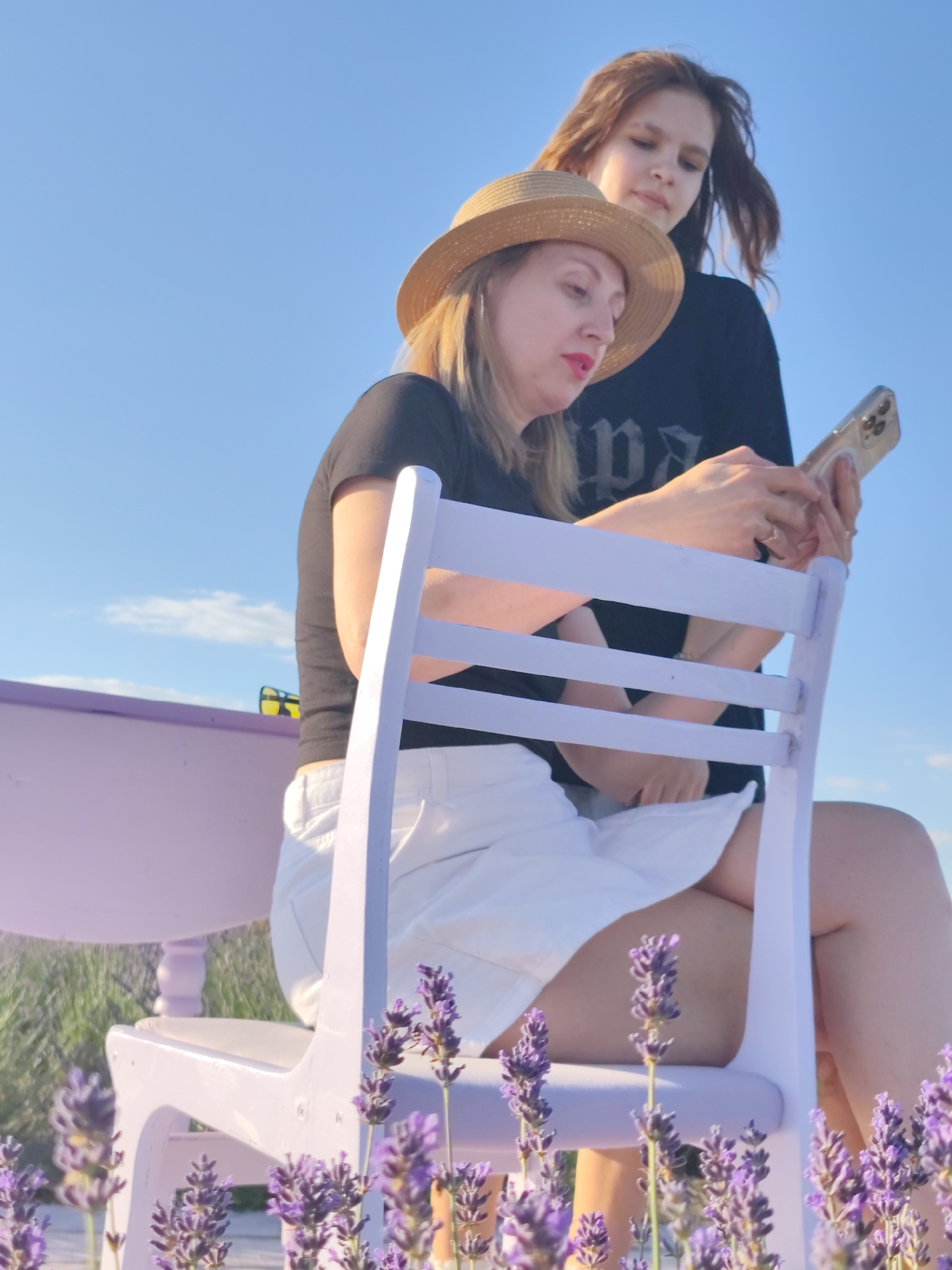 In Nechaiane, Ukraine, two people are seen outdoors with a clear blue sky above. A woman wearing a straw hat and a black t-shirt sits on a light purple chair, holding a mobile phone. She has bright pink lipstick on and appears to be looking at the phone. Behind her, another person, likely a younger female with long brown hair, stands and looks towards the woman with the phone. She is wearing a black t-shirt. A light purple bench is visible to the left of the frame, and a field of lavender is in the foreground, with some lavender sprigs reaching up in front of the chair. The scene suggests a relaxed, daytime setting, possibly in the late afternoon given the warm light. The overall atmosphere appears peaceful and candid. There is some faint text visible on the black t-shirt of the person standing behind, but it is not legible.