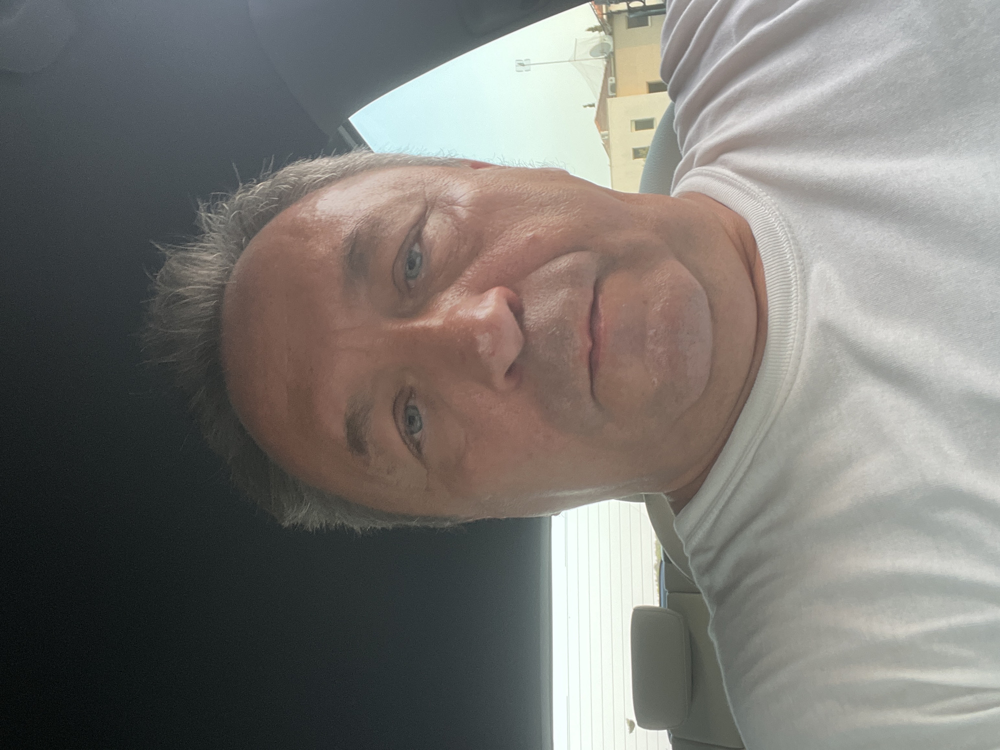 An outdoor, close-up selfie shows a middle-aged man with light skin, graying hair, and blue eyes. He is wearing a white t-shirt and is seated in the back of a car. The man has a neutral expression and is looking directly at the camera. The background is out of focus, but appears to be a residential area with buildings and a light blue sky. The image was taken in Parekklisia Community, Cyprus.