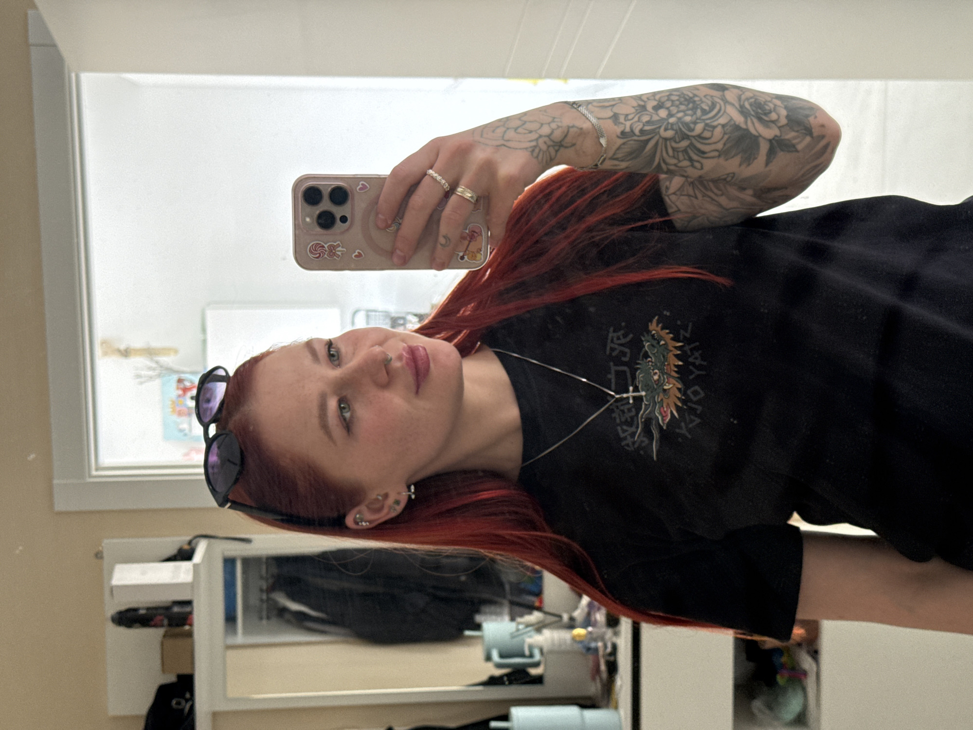 A young woman with red hair and a visible arm tattoo takes a selfie in a mirror. She wears a black t-shirt with a graphic design and has sunglasses perched on her head. She also has facial piercings and rings on her fingers. She is holding a cell phone to take the picture. The background shows a room interior. The room has a window and a shelving unit. No information indicates the image was taken in Rostov-on-Don, Russia.