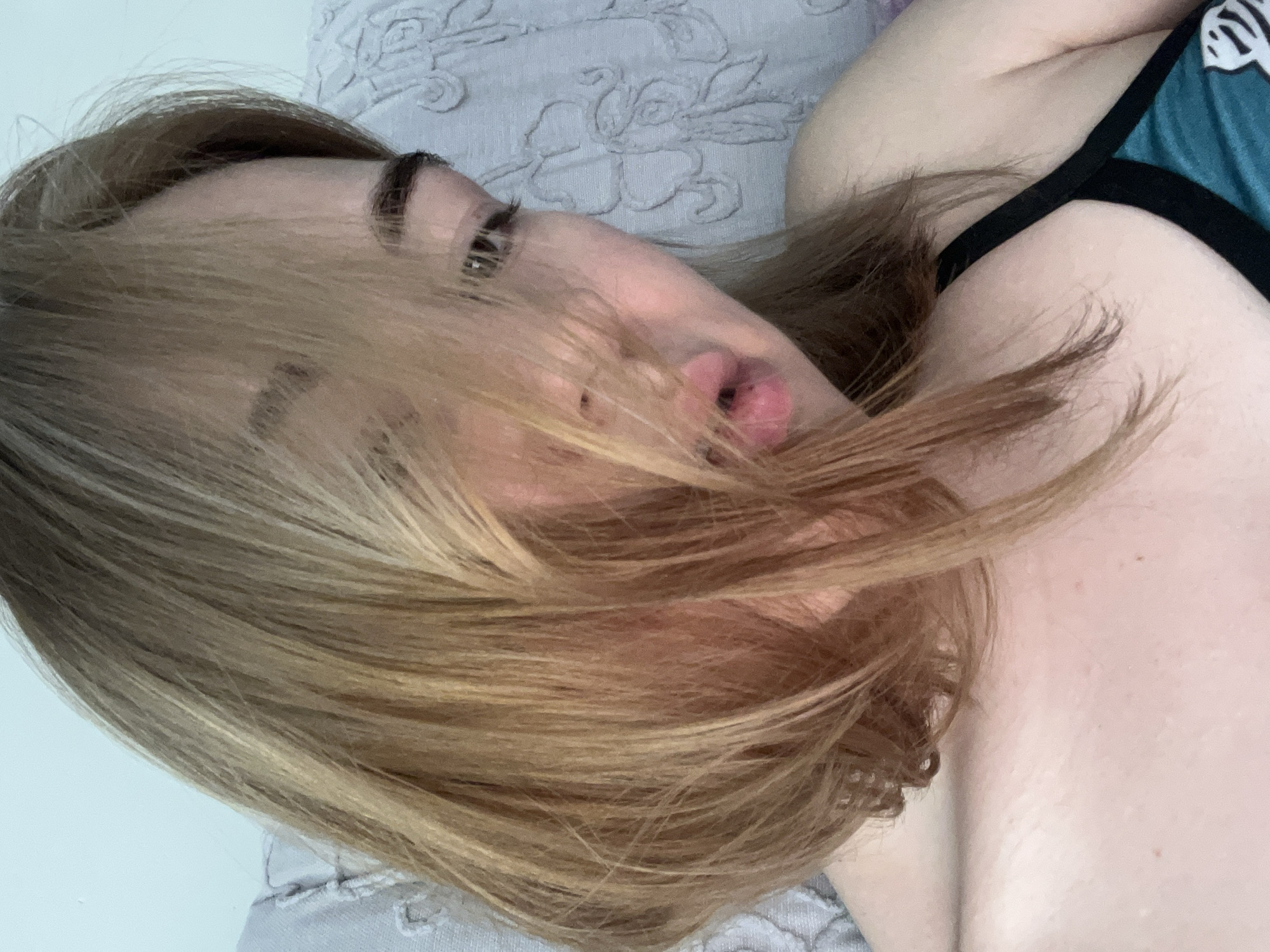 The scene, located in Boreti, Montenegro, depicts a close-up view of a female individual, likely a young adult, from the chest upwards. Her shoulder-length hair, colored light brown to blonde with lighter highlights, falls across and partially obscures her face, particularly the left side, forehead, and bridge of the nose. Her gaze is directed downwards and to the right within the frame. Her lips are slightly parted, revealing no teeth. The visible portion of her attire on the left side of the frame consists of a dark-colored strap and a teal-colored fabric section featuring a white, possibly floral or animal print pattern, consistent with a bra or swimwear top. The individual's skin tone appears fair. The background is composed of a light grey, textured fabric, possibly upholstery or bedding, with an embossed or embroidered floral or leaf motif visible on the right side and upper left. The lighting is soft and diffused, originating from the front-left, casting minimal shadows.