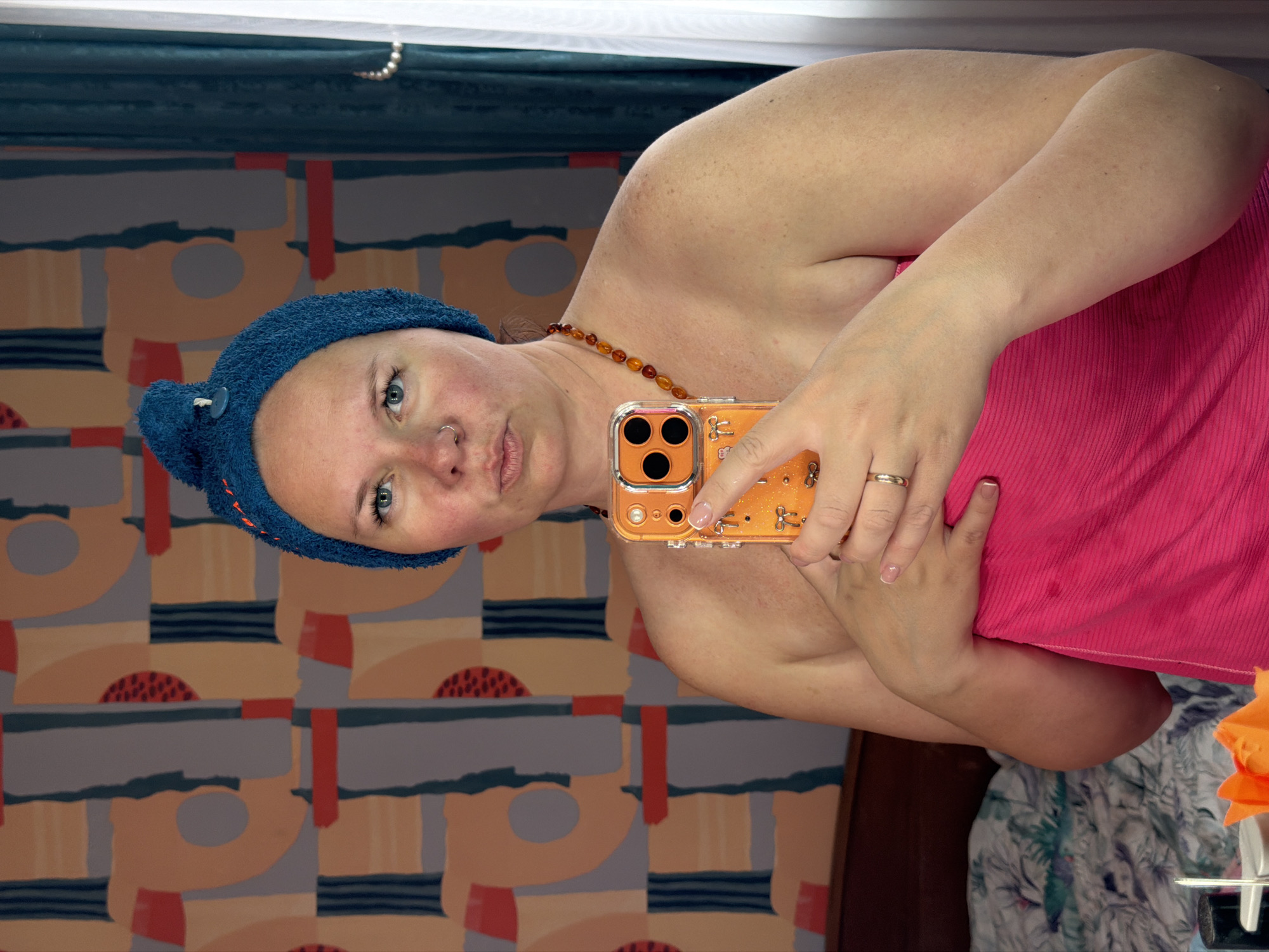The image shows a woman taking a selfie in a room with patterned wallpaper. The woman is wearing a blue towel on her head, a beaded necklace, and a pink top. She is holding an orange-colored phone with three circular elements on the back. A gold ring is visible on her finger. The background includes patterned wallpaper and a glimpse of what appears to be a white and blue floral fabric. The location in the city of Chaikovsky, Russia, is not discernible from the image.