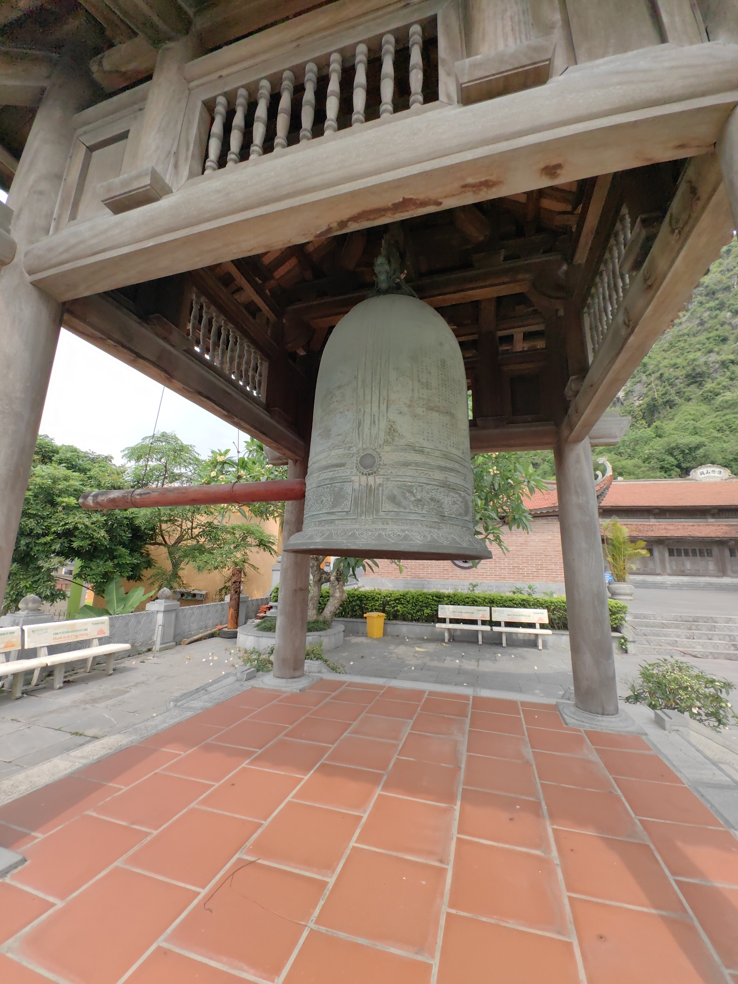 The image depicts a large bell hanging within a wooden structure in what appears to be an outdoor courtyard. The bell is prominently displayed, occupying a significant portion of the frame. The wooden structure surrounding the bell features intricate details, including balustrades and beams. The courtyard is tiled with rectangular terracotta-colored tiles. In the background, there are benches, greenery, and a building with a traditional red-tiled roof. Based on the prompt, the location is identified as Hong Gai Ward, Vietnam. There are no people visible in the image.