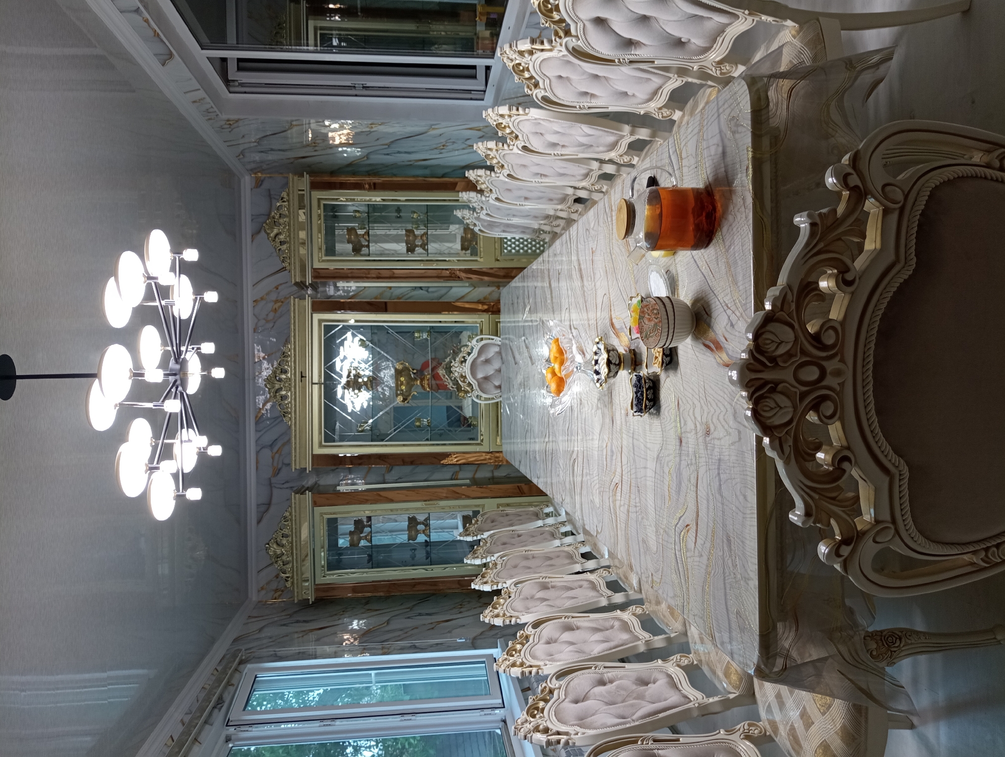 The image shows a formal dining room with a long table set for a meal. The room features ornate furniture, including a large table with a decorative tablecloth, and multiple upholstered chairs with carved wooden frames. On the table, there are several bowls containing fruit and other food items, as well as a glass teapot filled with amber-colored liquid. The walls are decorated with a marble-patterned wallpaper, and tall display cabinets with glass doors line the sides of the room. A modern chandelier hangs from the ceiling. The overall impression is one of luxury and formality. This dining room is located in Shymkent, Kazakhstan.