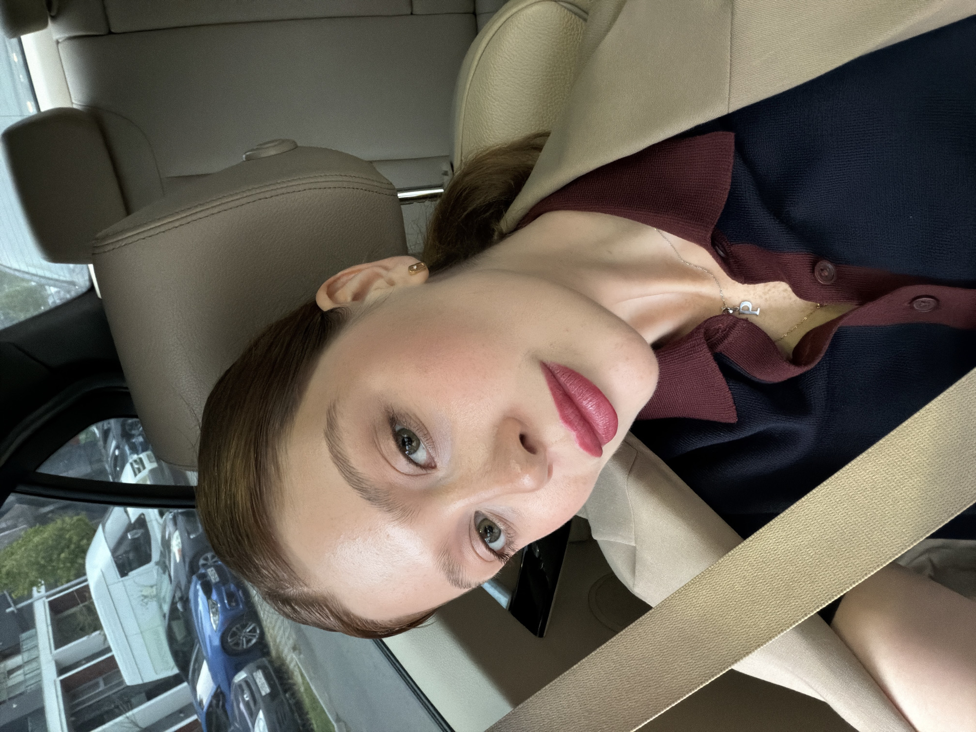A close-up portrait shows a young woman seated in what appears to be the passenger seat of a car. She looks directly at the camera with a neutral expression. Her hair is pulled back, revealing a small gold ear cuff on her right ear. She has green eyes, defined eyebrows, and is wearing dark pink lipstick and subtle cheek makeup.
She is dressed in a collared top or cardigan featuring dark blue and burgundy sections, layered under a light beige blazer or coat. A silver necklace with a pendant resembling the letter 'P' is visible around her neck. A beige seatbelt crosses diagonally over her chest.
The car's interior features beige leather seats and headrests. Through the car window, an urban environment is visible, with other vehicles (including a prominent blue car), buildings, and some greenery suggesting a city street. The lighting is soft, indicating daytime, possibly overcast. The location is specified as Tbilisi, Georgia.