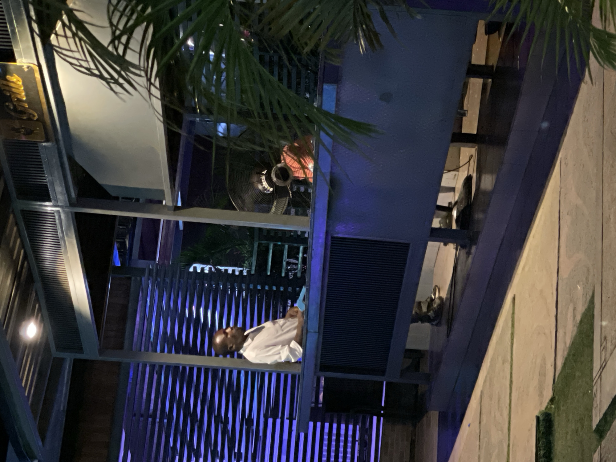 A man stands on an elevated outdoor platform under a palm tree. He is dressed in a white shirt and looking at a handheld device. The platform is lit with blue and purple lighting, and it appears to be part of an outdoor dining or entertainment area, potentially associated with "Grills," as suggested by signage. A fan is visible in the background.