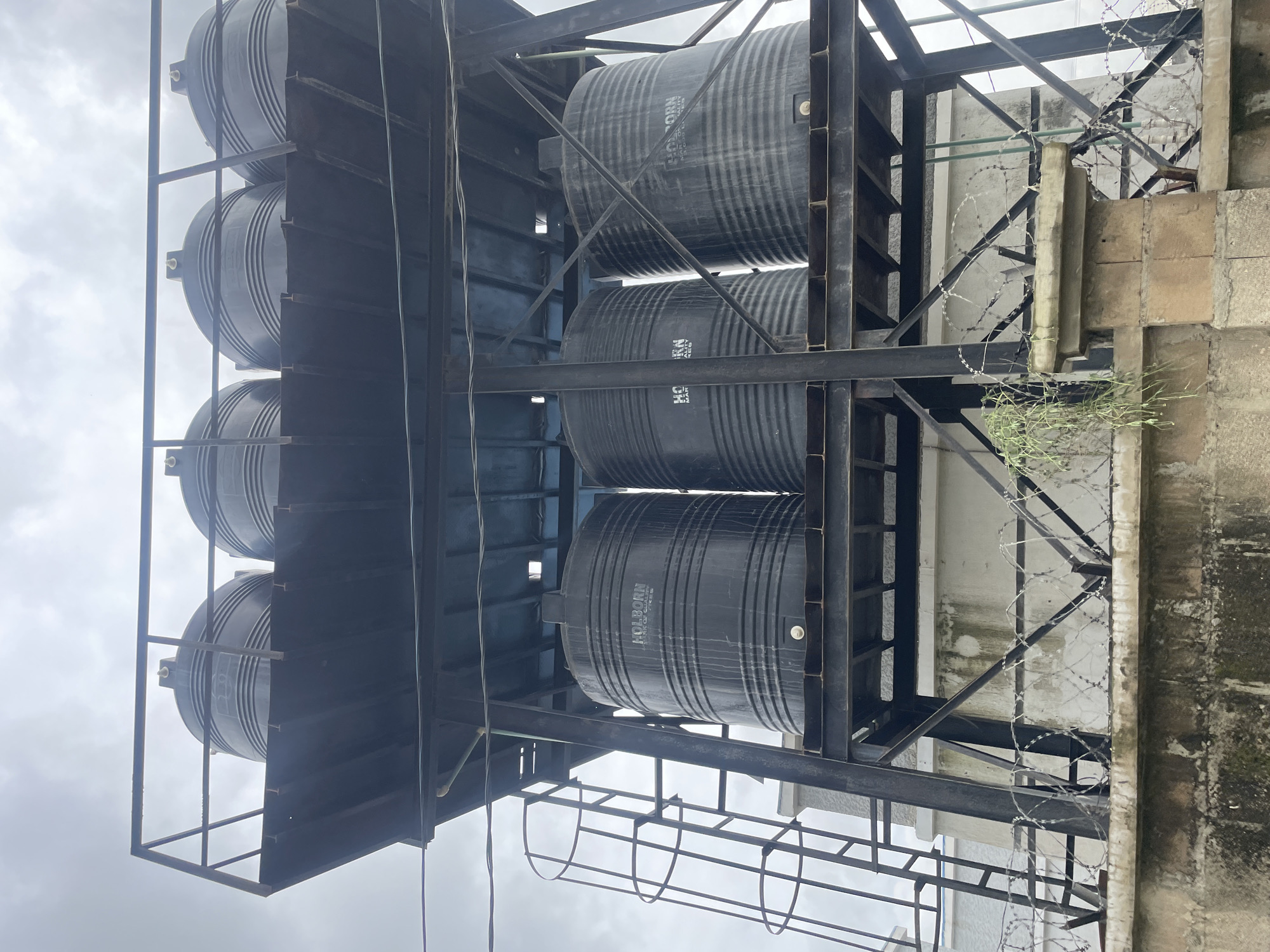 A low-angle, full-shot photograph shows a metal water tower holding four large, black, ribbed water tanks. The tanks are arranged in two rows of two on a black metal platform, with the front two tanks being closer and larger in the frame. The platform is supported by a dark metal structure with diagonal bracing. A metal ladder with a circular safety cage is attached to the left side of the structure, leading up to the platform. The tanks have the brand name "HOLBORN" visible on their sides. Below the water tower, a concrete wall topped with barbed wire is visible, with some vegetation growing on it and hanging from the water tower structure. The sky above is overcast and cloudy. The location is identified as Unknown, Nigeria.