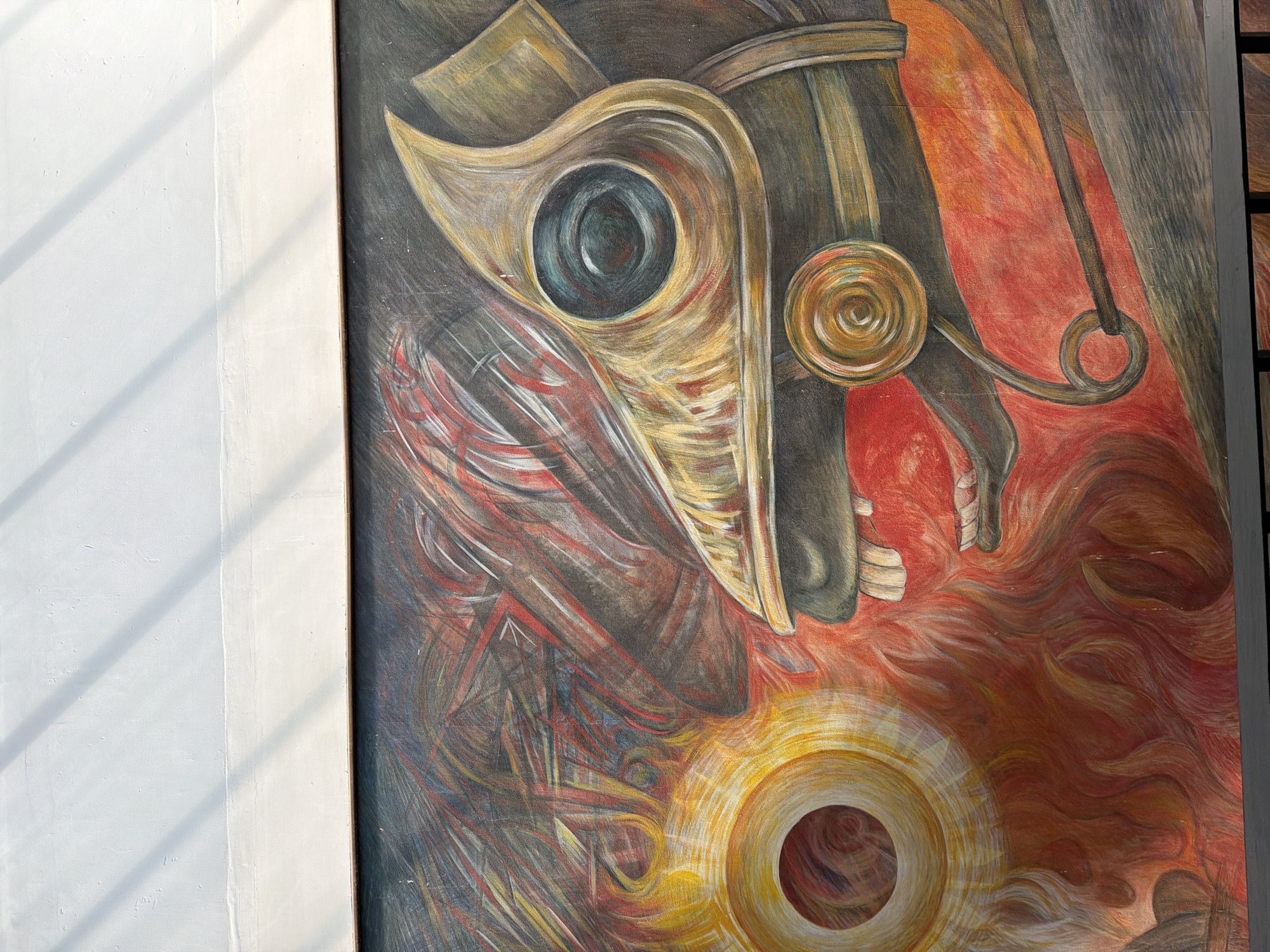The image shows a section of a mural. The mural depicts an abstract figure resembling a horse's head or a mask, with metallic and swirling elements in shades of gold, black, red, and grey. The mural features a dark, central "eye" shape with concentric rings, and other circular and spiraled forms. The mural is attached to a white wall, likely in a public space, perhaps located in San Andrés Cholula, Mexico.