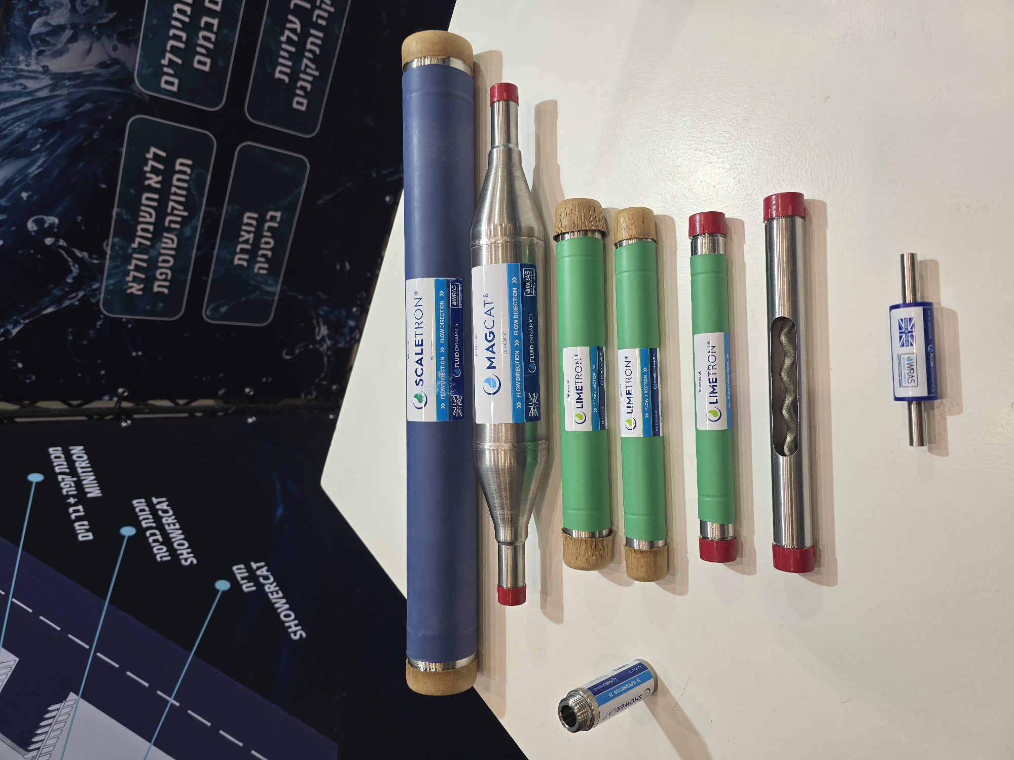 The image shows a display of various water treatment devices on a white surface. The devices include Scaletron, Magcat, and Limetron models. Labels indicating "Flow Direction" and the WRAS (Water Regulations Advisory Scheme) certification are visible on the devices. A small device labeled "Showercat" is also present. The image also contains a partial view of a sign in Hebrew with English translations indicating the benefits of the products, such as "mineral in the water," "without electricity and without routine maintenance," and applications like "coffee machine + bar water Minitron" and "dishwasher Showercat".
There is no visible information to connect the products to Tel-Aviv, Israel.