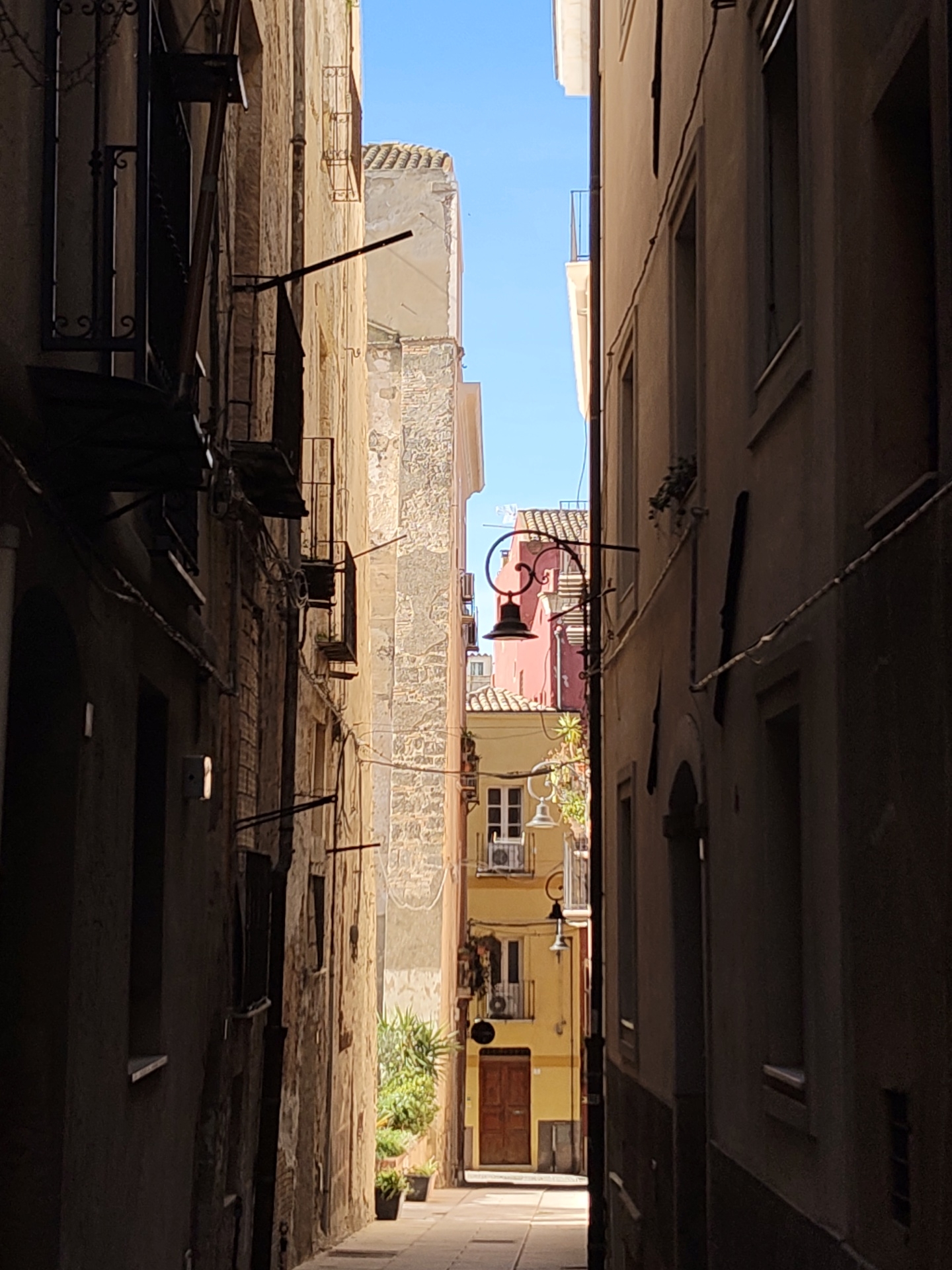 A narrow street in Cagliari, Italy, is framed by aged buildings. The left side features a dark, weathered stone building with iron balconies and window bars. The building on the right is lighter in color, with evenly spaced windows. In the distance, the street opens to a view of colorful buildings, including yellow and pink facades, visible under a clear blue sky. Architectural details such as a wrought-iron street lamp and potted plants add to the scene. The street is paved with light-colored stones.