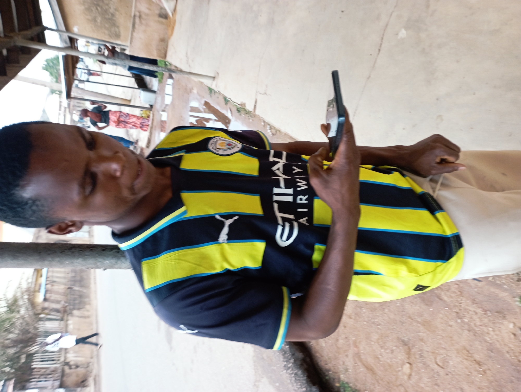 A man is looking at his phone on a partially shaded, paved area in Jos, Nigeria. The man is wearing a striped, yellow and black soccer jersey and khaki pants. The phone is black. In the background are buildings, trees and people.